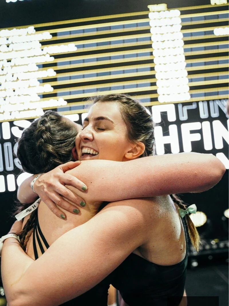Two women hugging each other with smiles after a fitness event, in front of a digital display board showing event information.