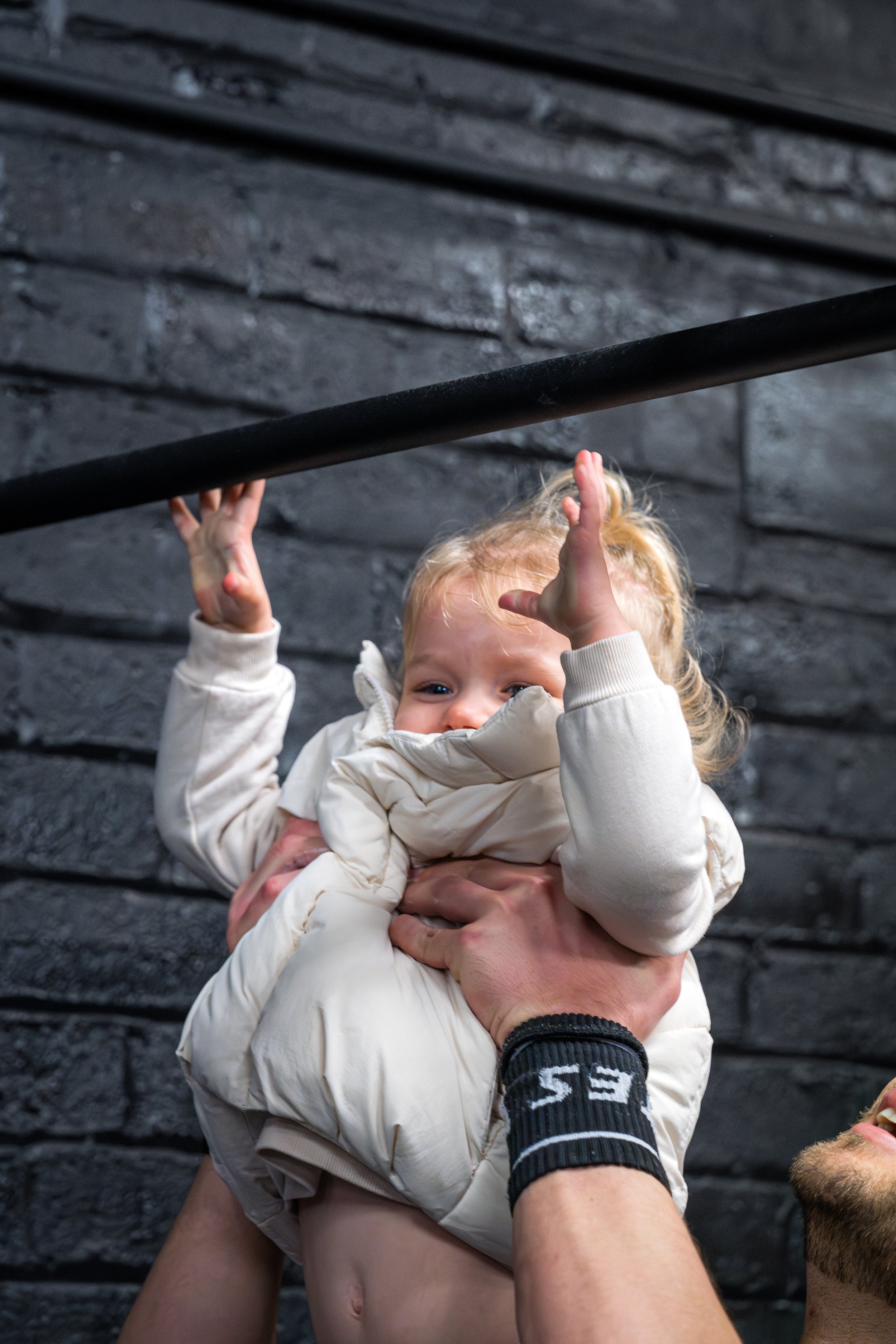 A person with a wristband holding a young girl with blonde hair and light-colored clothing. The girl is smiling and reaching up with one hand. They are in front of a dark, textured brick wall.