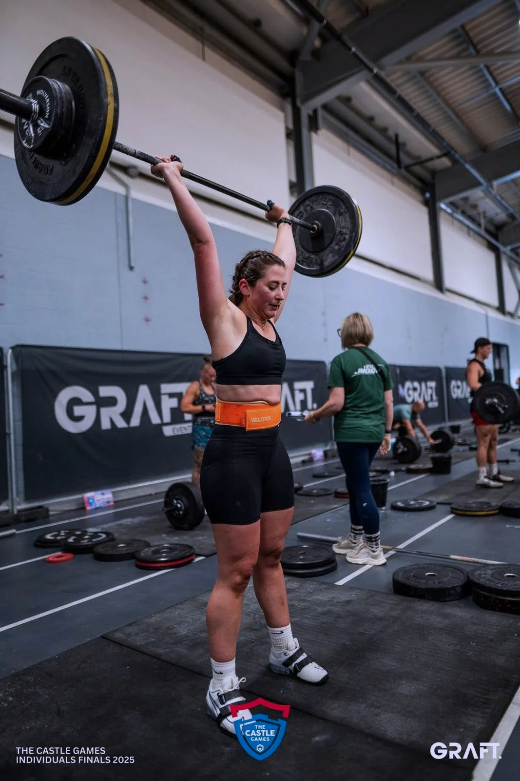 A woman participating in a weightlifting competition, lifting a barbell overhead at the Graft Castle Games, individual finals 2025, in a large indoor gym.