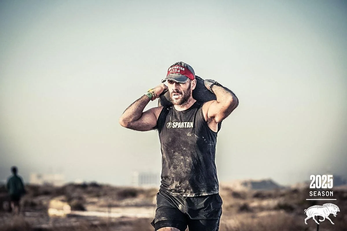 A man wearing a red cap, a black sleeveless shirt with 'SPARTAN' written on it, and black shorts, carrying a large bag on his shoulders during a rugged outdoor race, with a hazy sky and rocky landscape in the background.