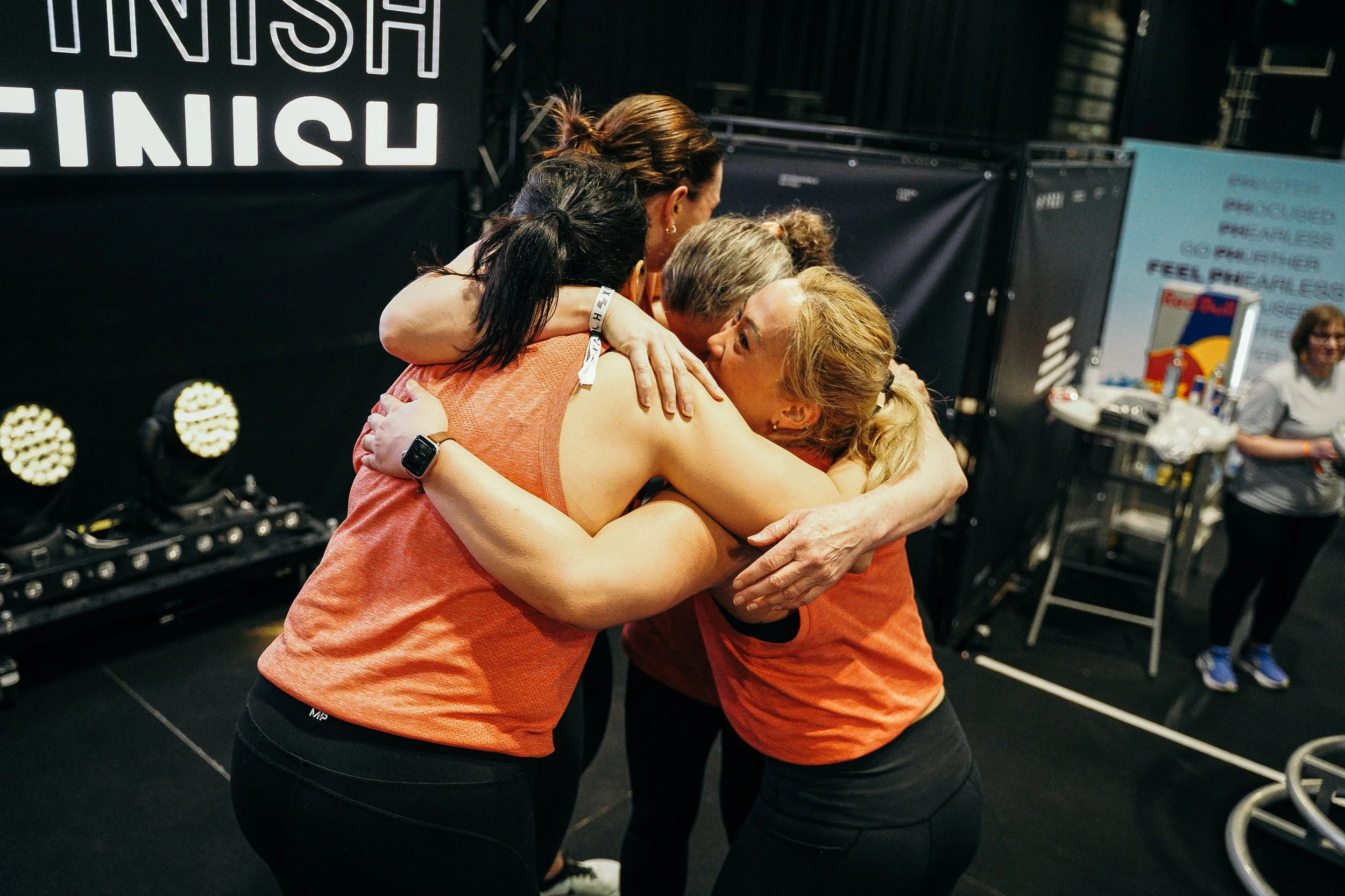 A group of women cheer and hug in a gym or fitness center after completing a race, with a large 'Finish' sign in the background.