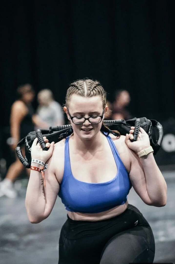 A woman in athletic attire carrying a weighted bag on her shoulders during a workout or competition, with others in the background.