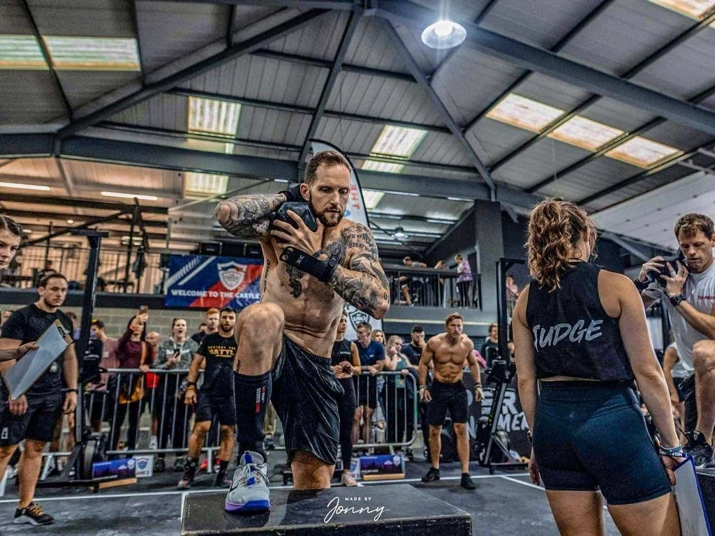A muscular man with tattoos on his arms and chest preparing to do a workout with a medicine ball in a gym, surrounded by other people, some of whom are working out, taking photos, and watching.