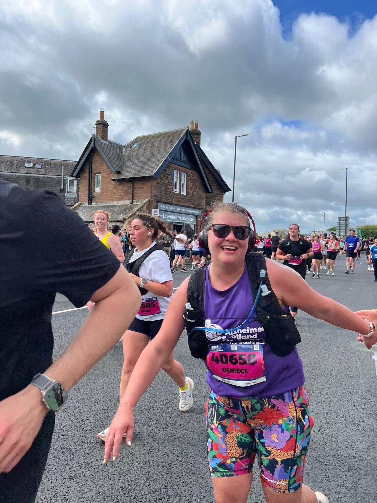 A woman smiling, wearing sunglasses and headphones, participating in a marathon, surrounded by other runners in an outdoor setting with cloudy sky and a brick house in the background.