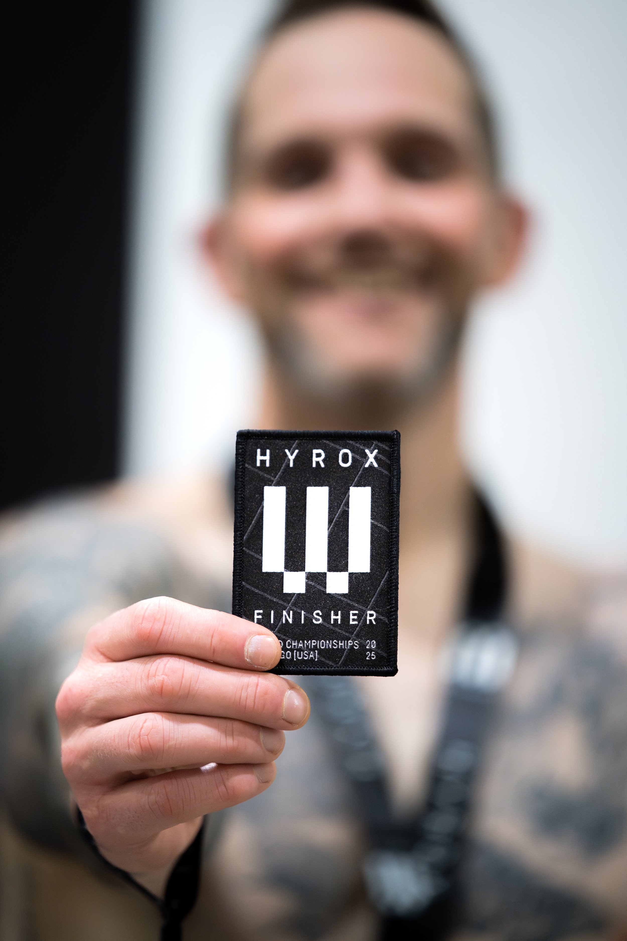Person holding a black finisher patch with white text and design, smiling, blurred background.
