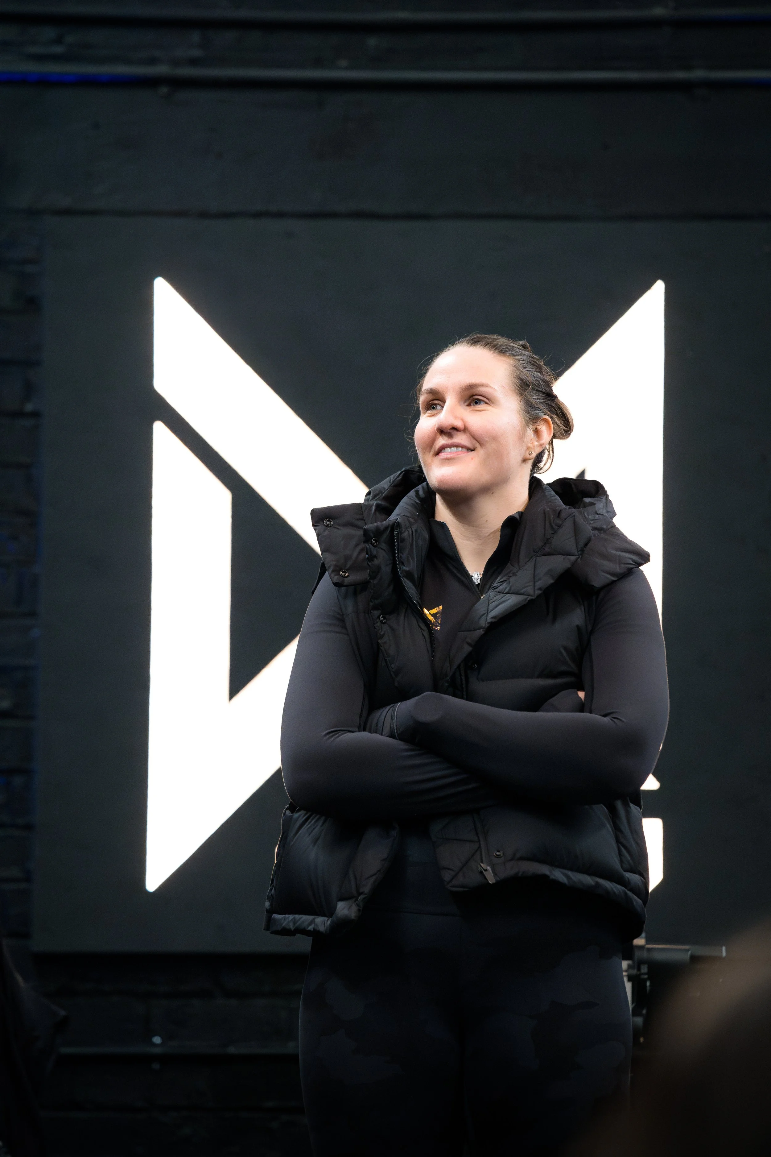 A woman in athletic clothing with arms crossed, standing in front of a dark wall featuring a large, illuminated geometric design.