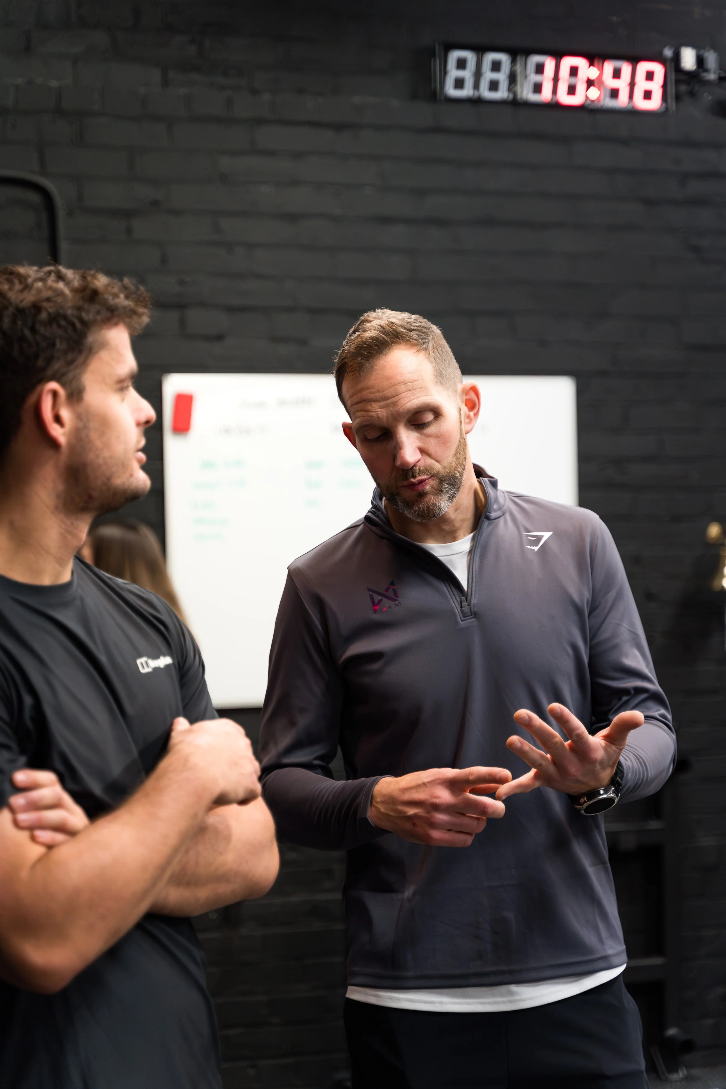 Two men engaging in conversation in a gym, with a digital clock displaying 10:48 in the background.
