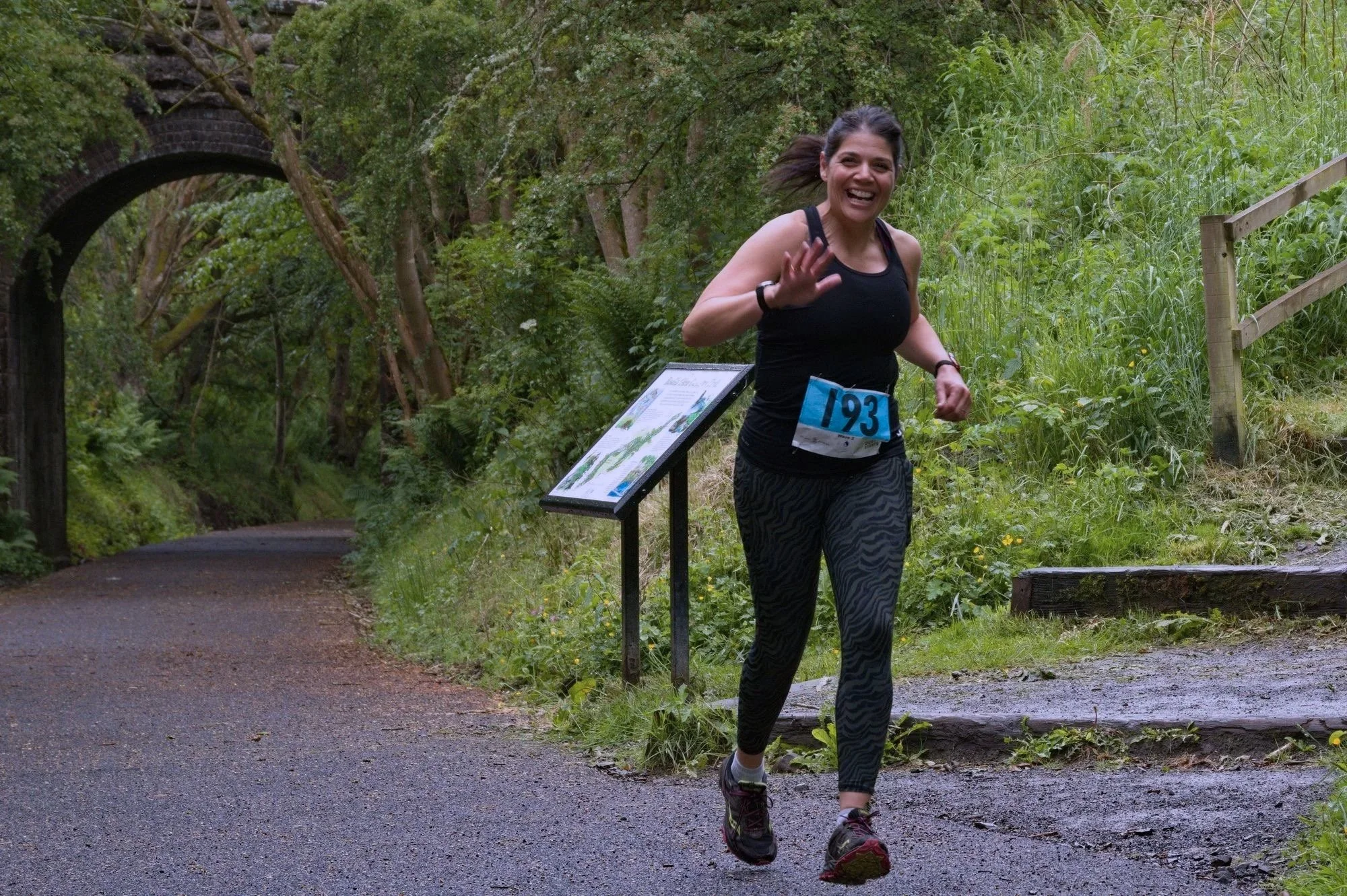 A woman running on a forest trail during a race, wearing a bib number 193, black tank top, patterned leggings, and smiling and waving at the camera.