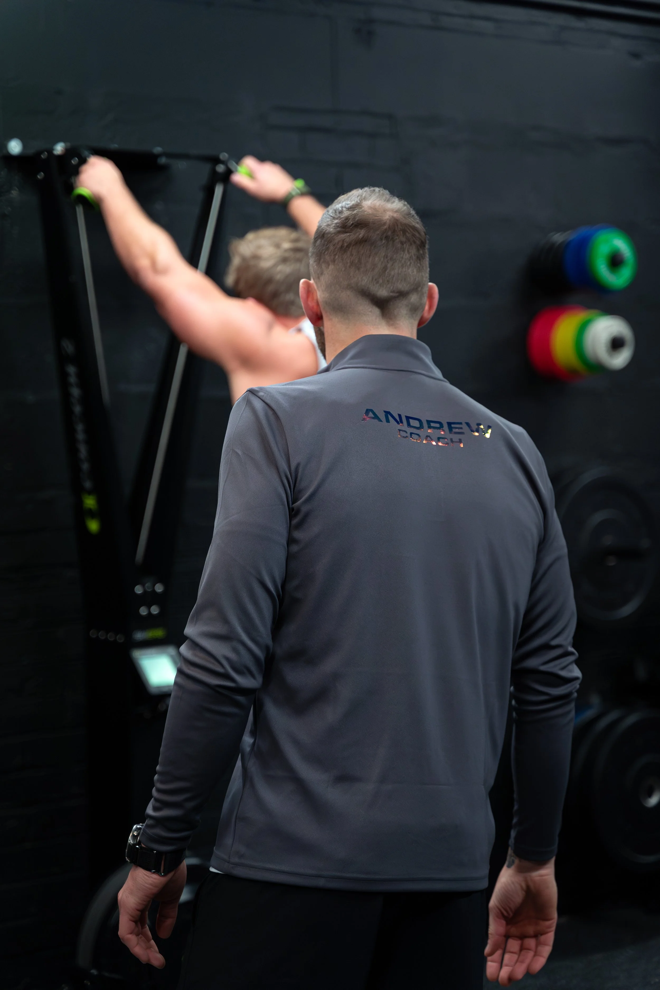 A fitness trainer observing a male client performing exercises on a wall-mounted exercise rack in a gym.