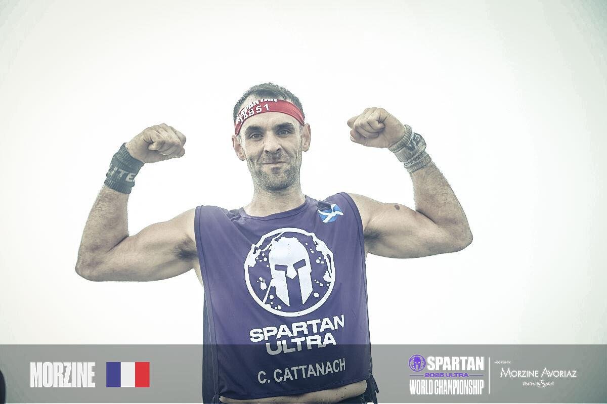 A man wearing a blue Spartans Ultra tank top and red headband flexes his biceps, smiling at the camera, against a neutral background.