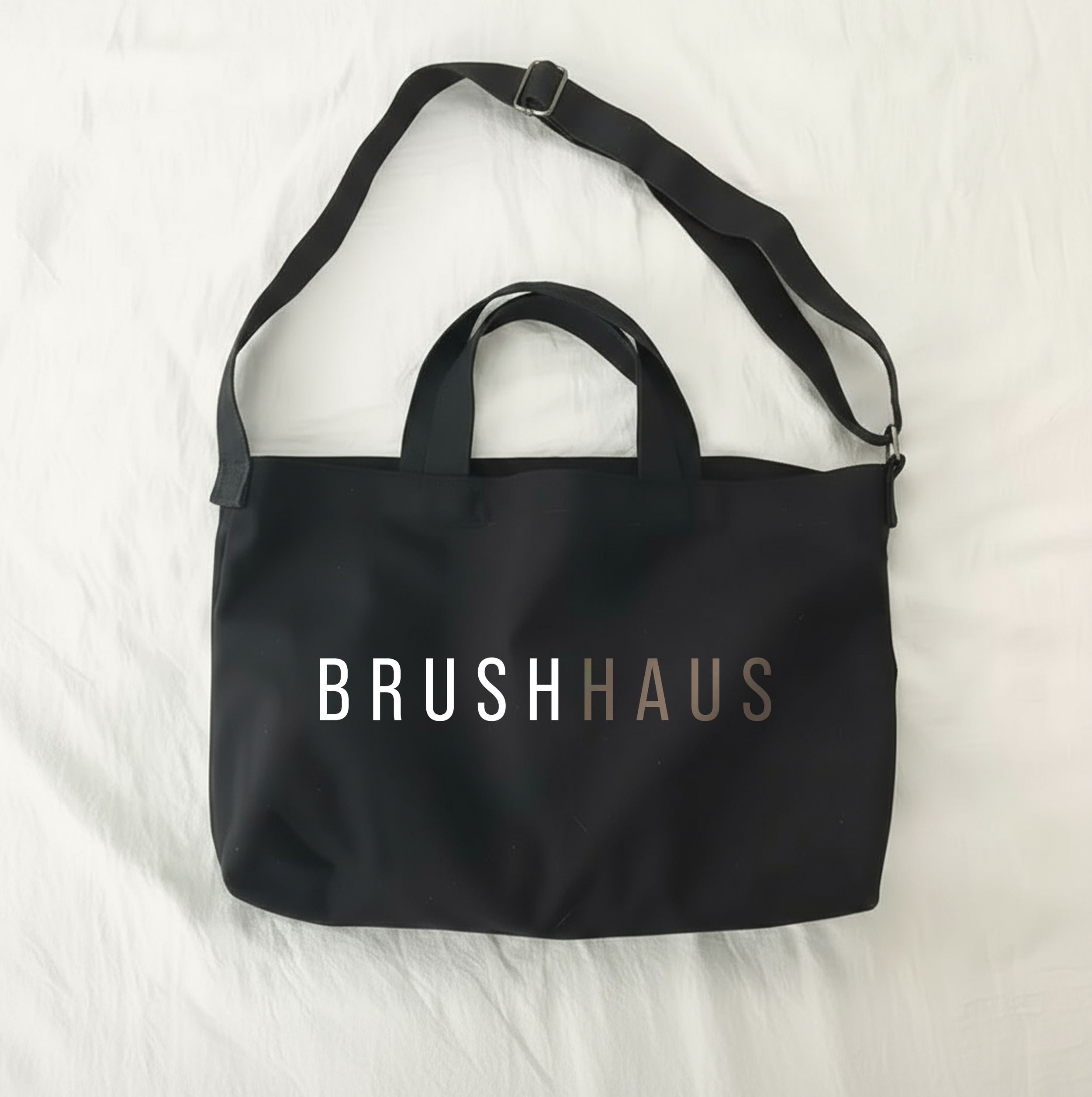 A black tote bag with handles and an adjustable shoulder strap on a white surface. The bag displays the text 'BRUSH HAUS' in white and gray letters.