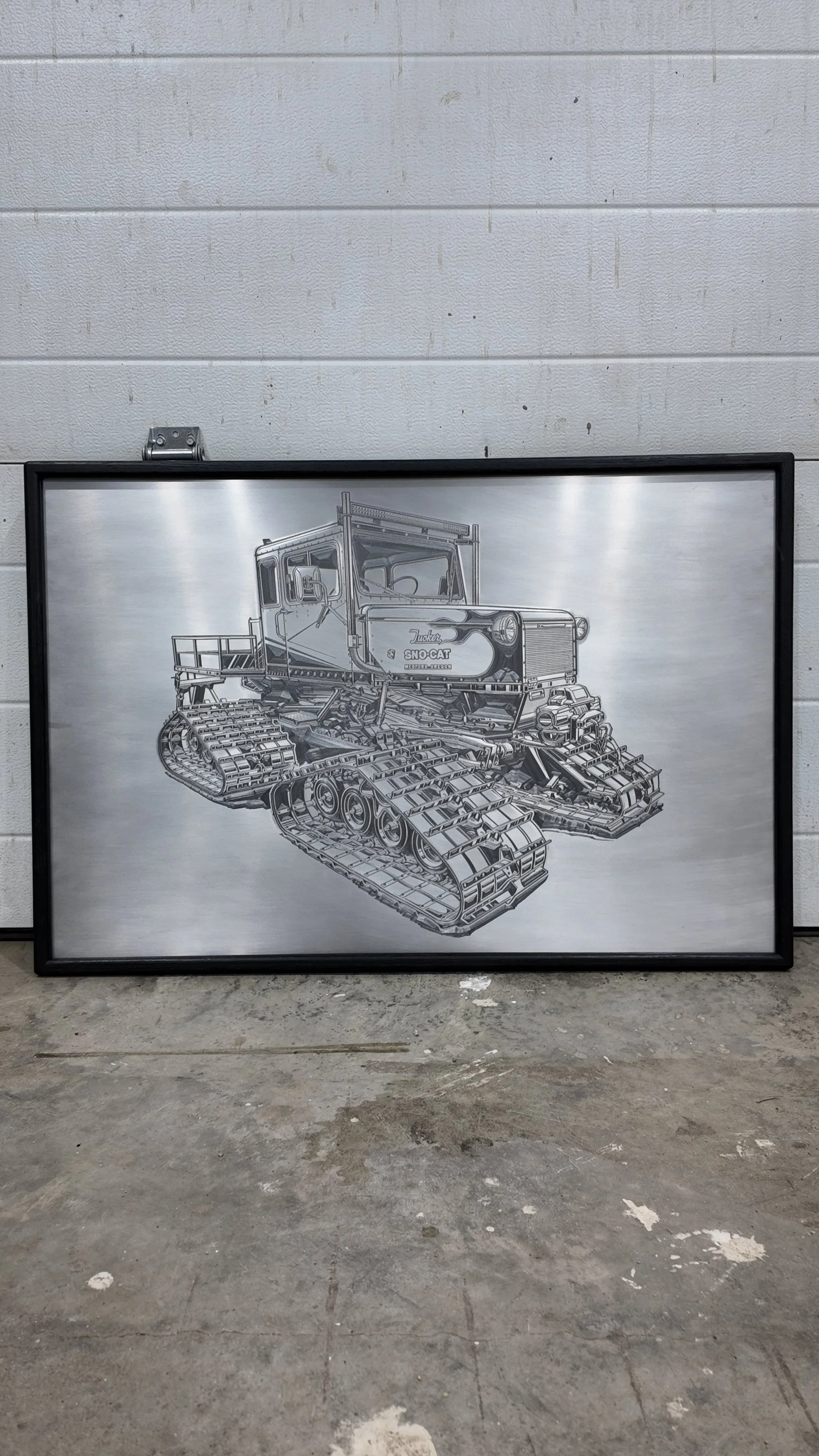 Framed black-and-white technical drawing of a tracked vehicle, leaning against a wall in an industrial setting.