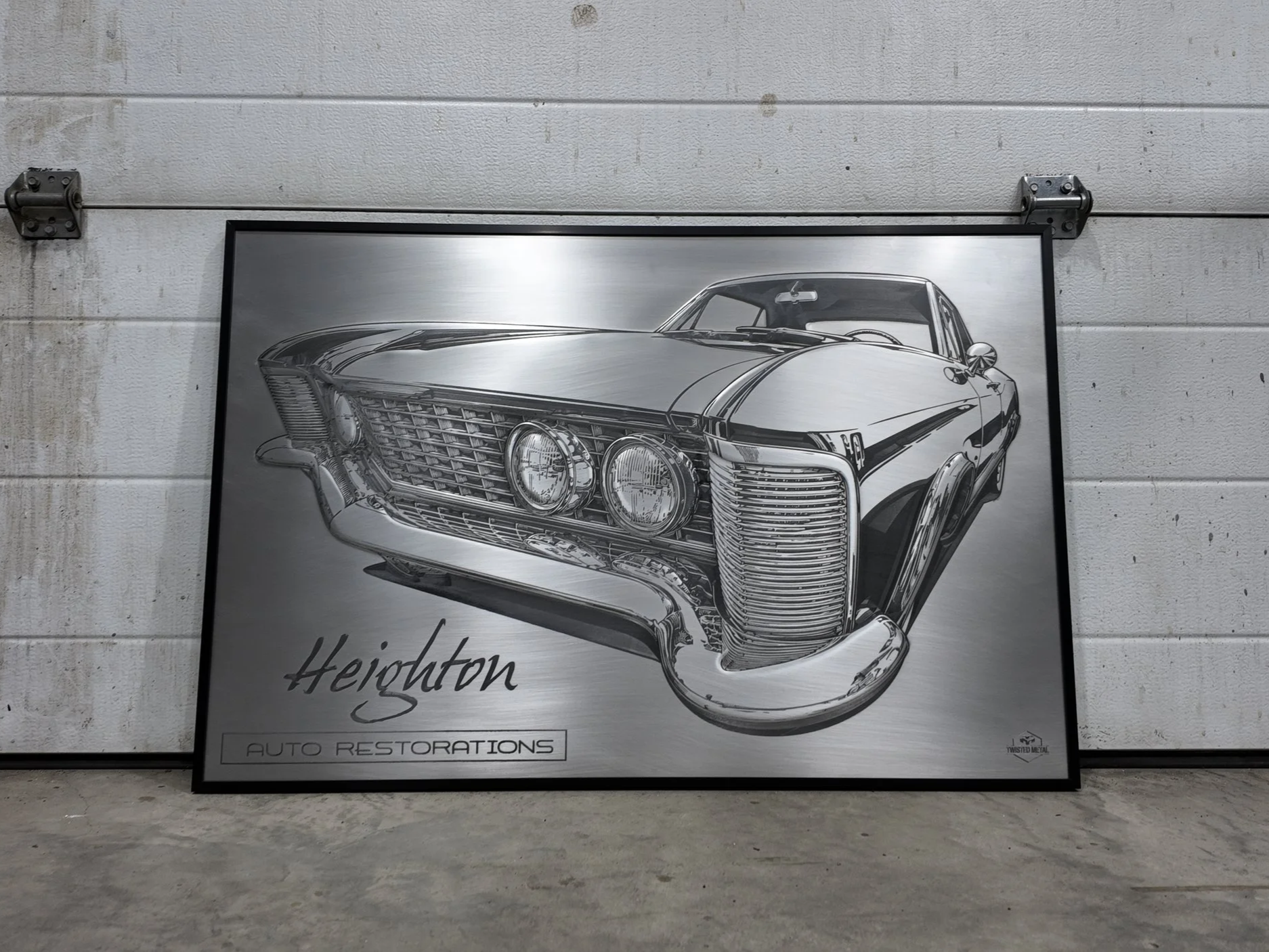 A black and white framed poster features a detailed illustration of a vintage car with prominent front grille and dual headlights. The poster has the text 'Heights' and 'Auto Restorations' on it. The background shows a plain, textured wall with metal hardware.