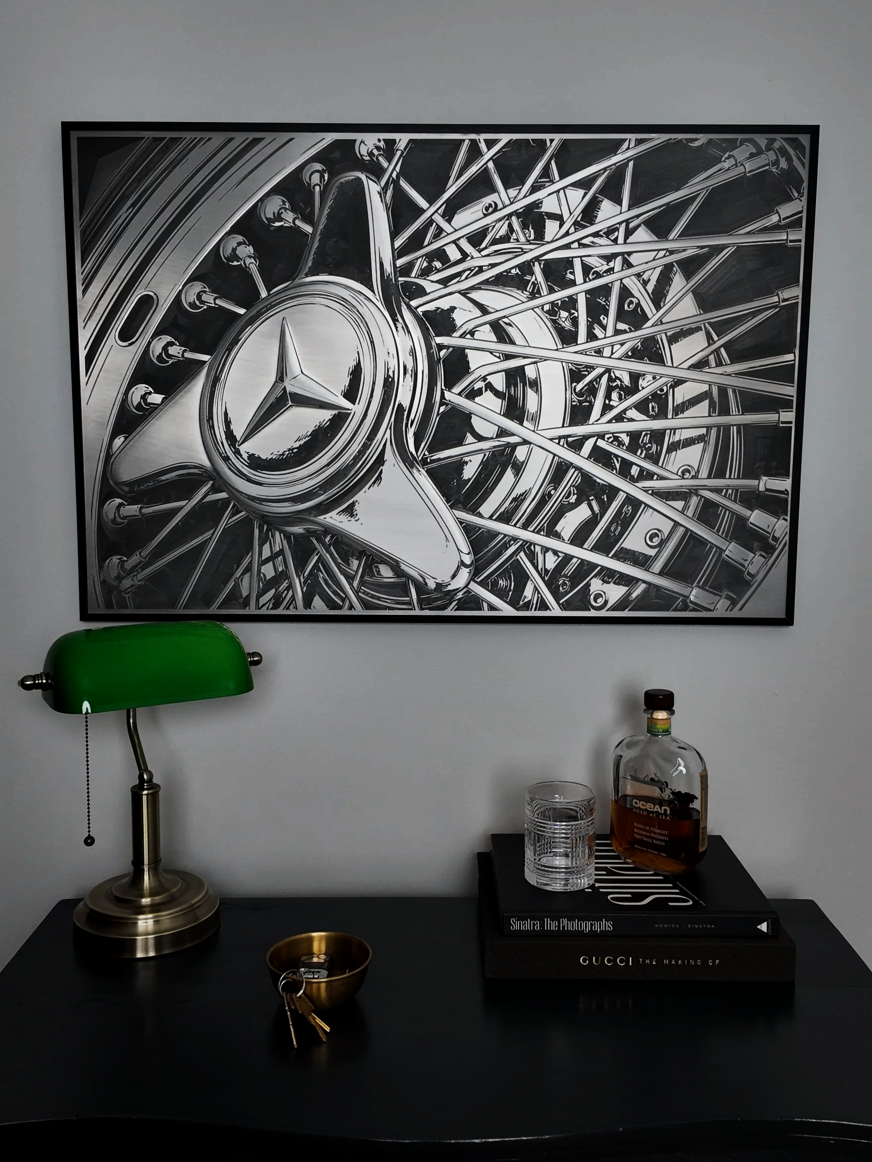 A black and white artwork of a Mercedes-Benz logo on a car wheel hubcap with multiple spokes. The artwork is hanging on a plain wall.