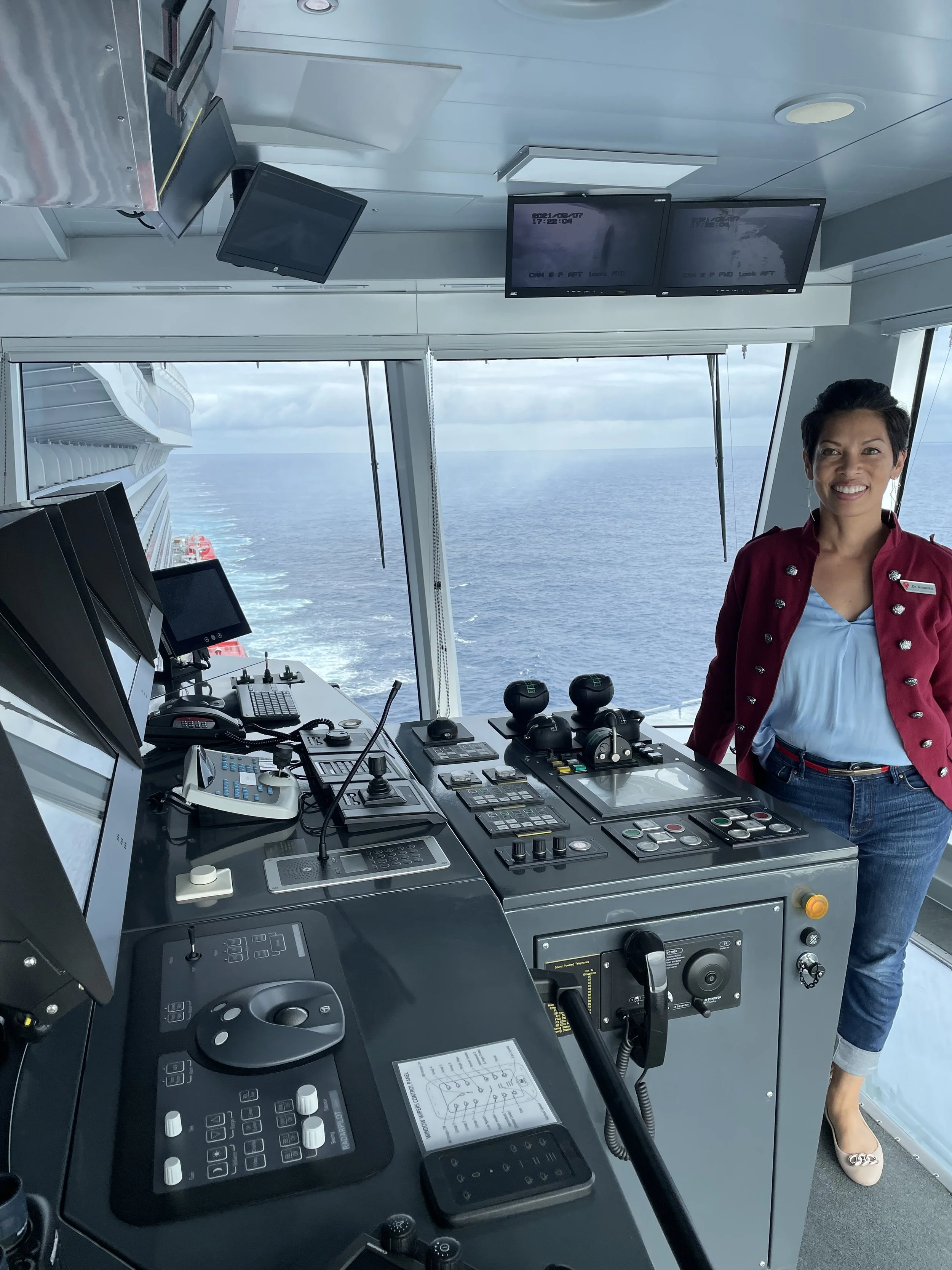 A smiling woman standing at the navigation control panel on a cruise ship bridge with the ocean view through large windows behind her.