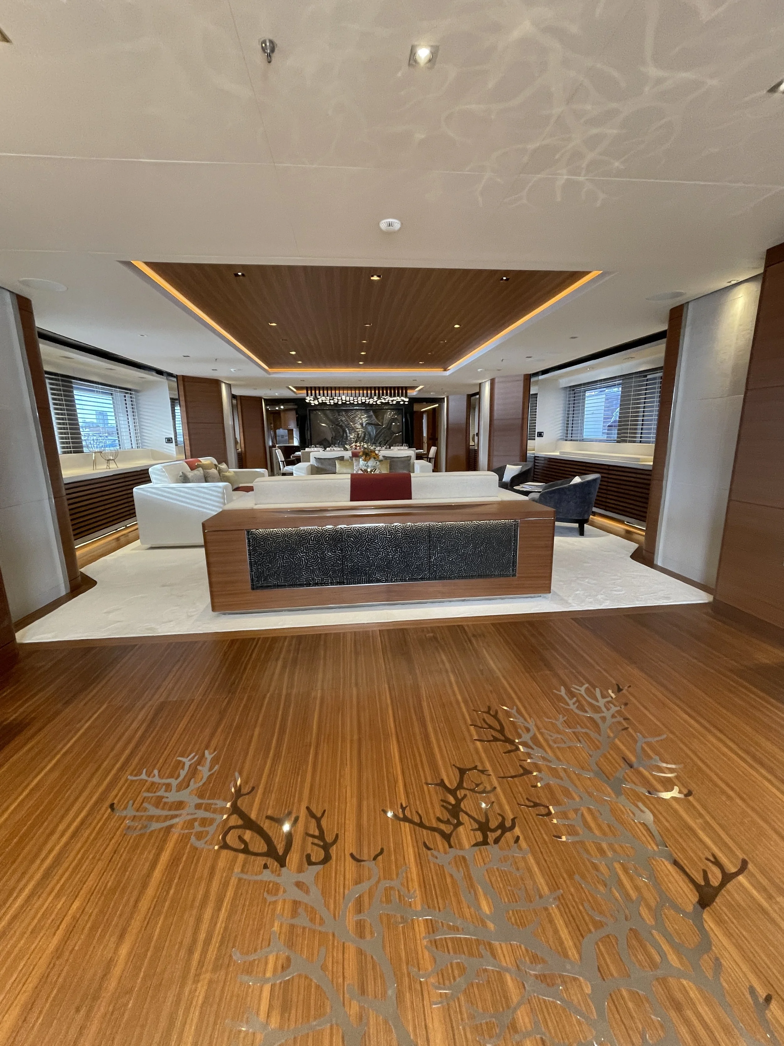 Elegant hotel lobby with wooden floors, modern furniture, and large windows. Centered seating area with white sofas and armchairs, decorative lighting on ceiling, and artwork on walls.