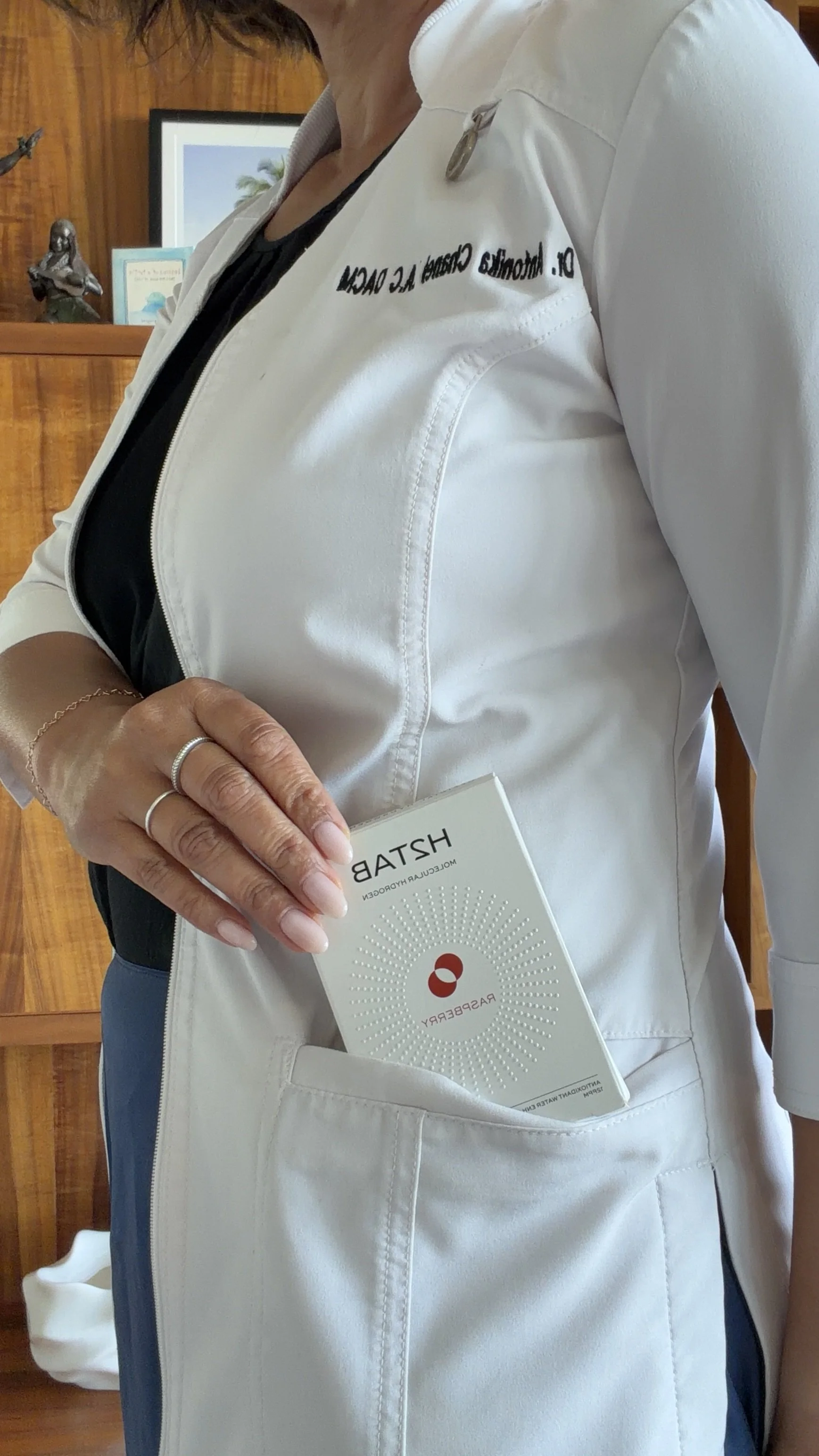 Close-up of a person wearing a white medical coat, holding a pamphlet, with part of the hand and coat in view. The background shows wooden furniture and framed pictures.