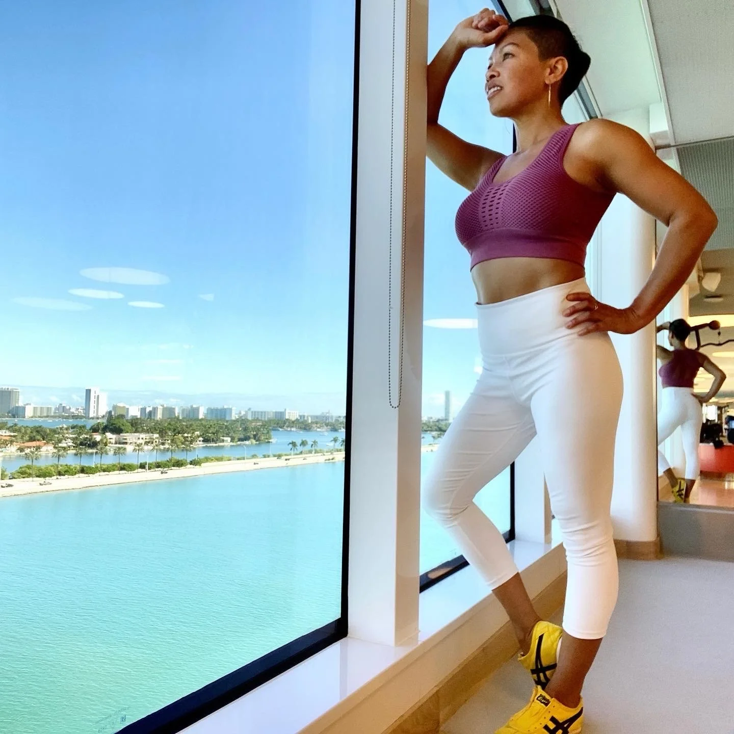 A woman in workout clothes standing by a large window overlooking a body of water with a city skyline in the distance. She is posing with one hand on her hip and the other resting on her head, looking outside.