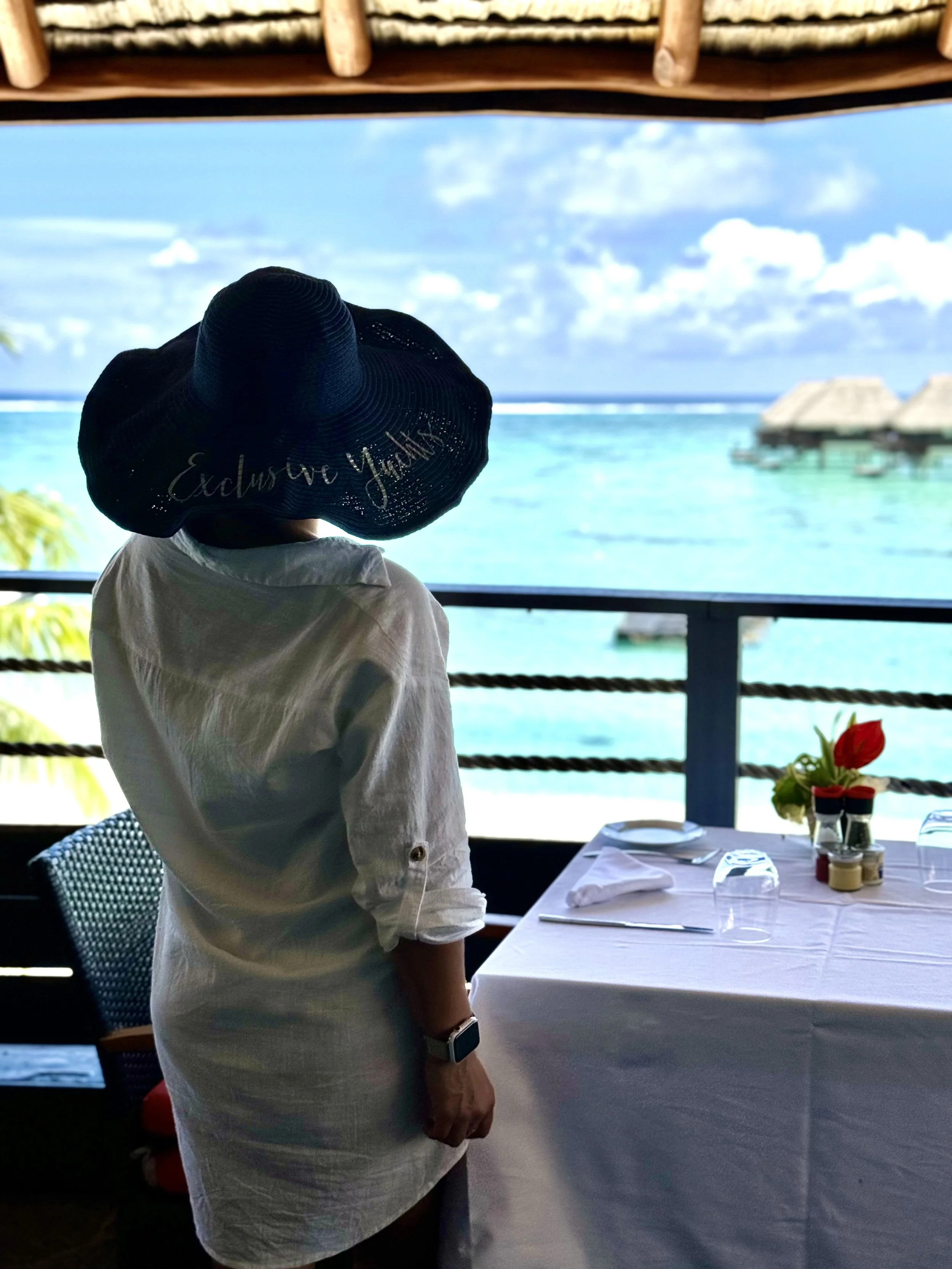 Woman standing inside a restaurant, wearing a large sun hat with "Exclusively You" embroidered on it, looking out at the ocean and overwater bungalows.