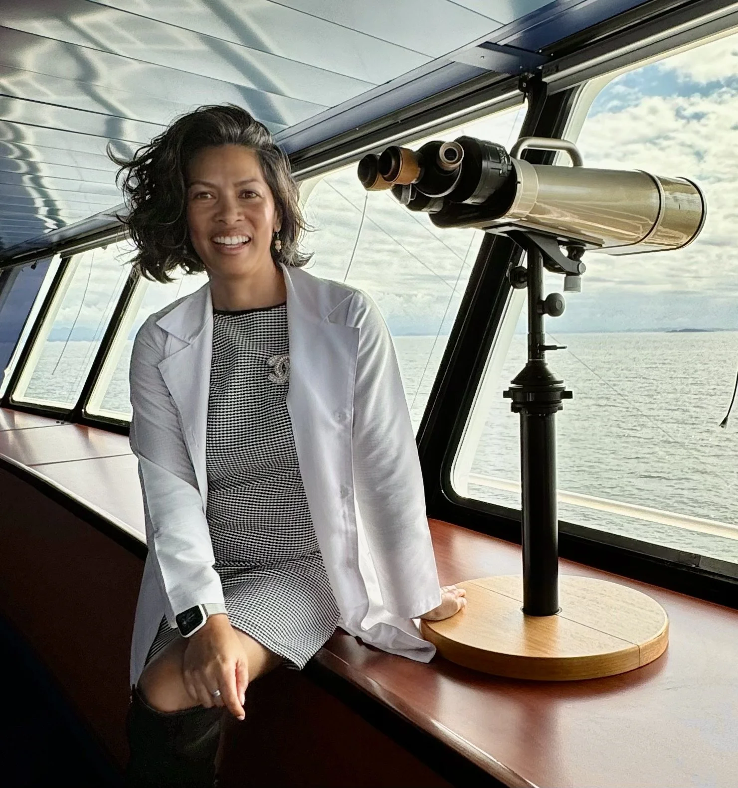 Woman in a white lab coat sitting on a ship's deck near a telescope, with ocean and sky visible through the windows.