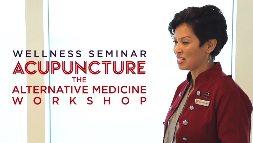 A woman standing indoors near a window, wearing a red blazer with name tags, participating in a wellness seminar about acupuncture, alternative medicine, and the workshop.
