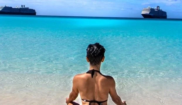 A woman sitting on the sandy beach facing the turquoise ocean with cruise ships in the distance.