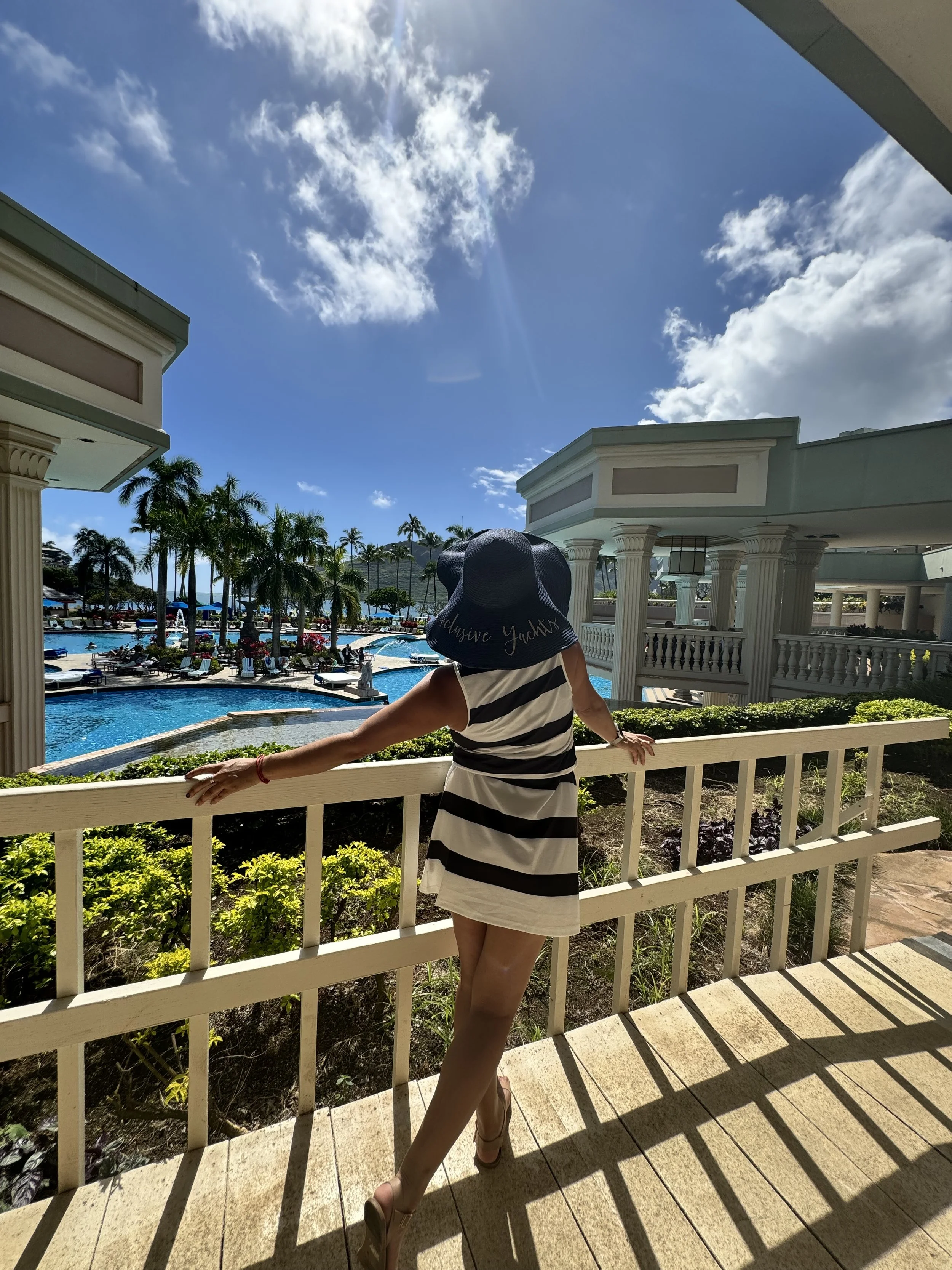 Woman in a striped dress and large sun hat standing on a balcony overlooking a resort pool area with palm trees and blue sky.