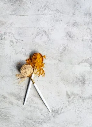 Two white sprigs with spoons containing turmeric and ginger powder on a gray textured background.