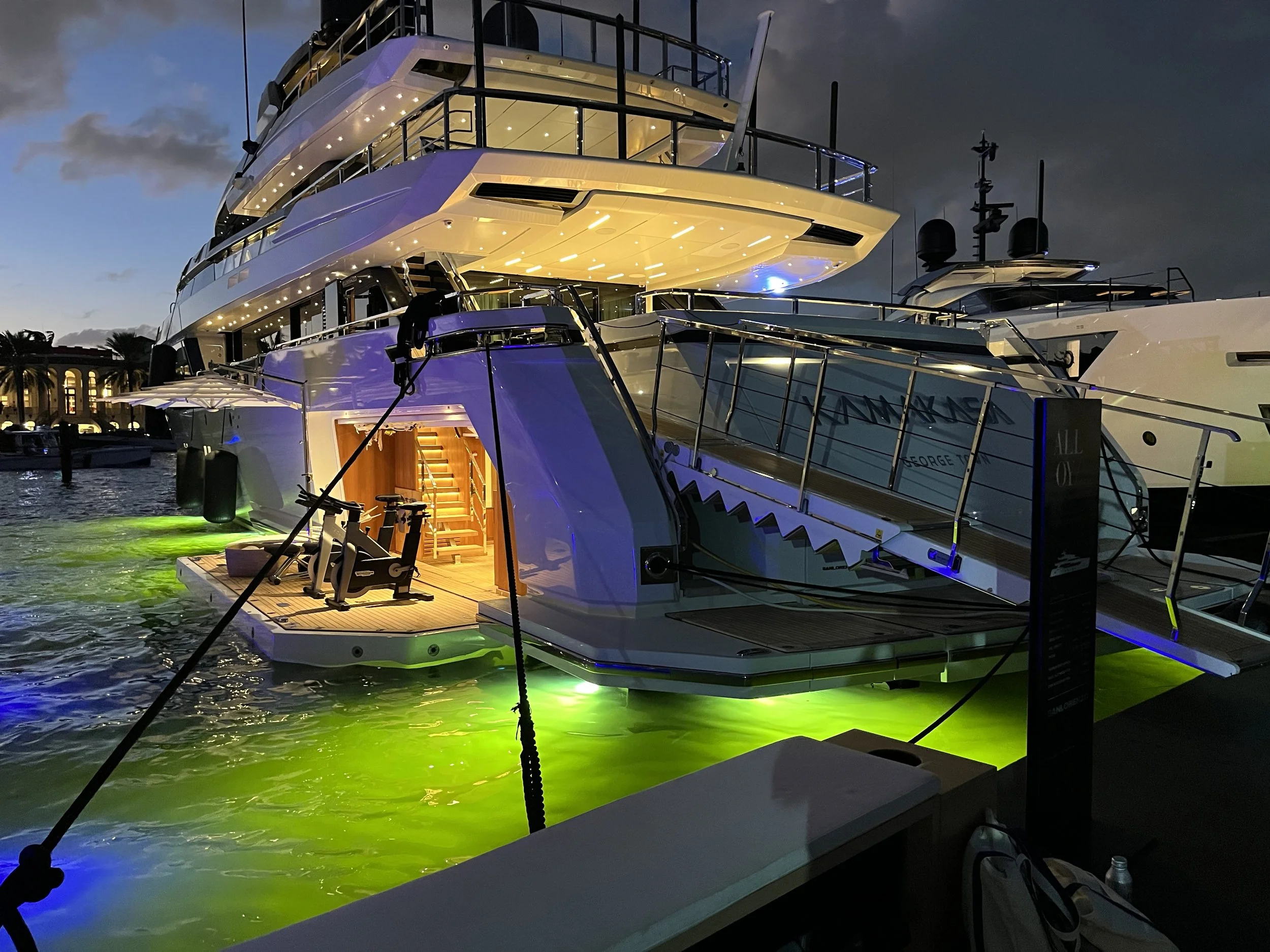 Large luxury yacht docked at a marina during evening, with green underwater lights, illuminated interior, and exercise equipment on the lower deck.
