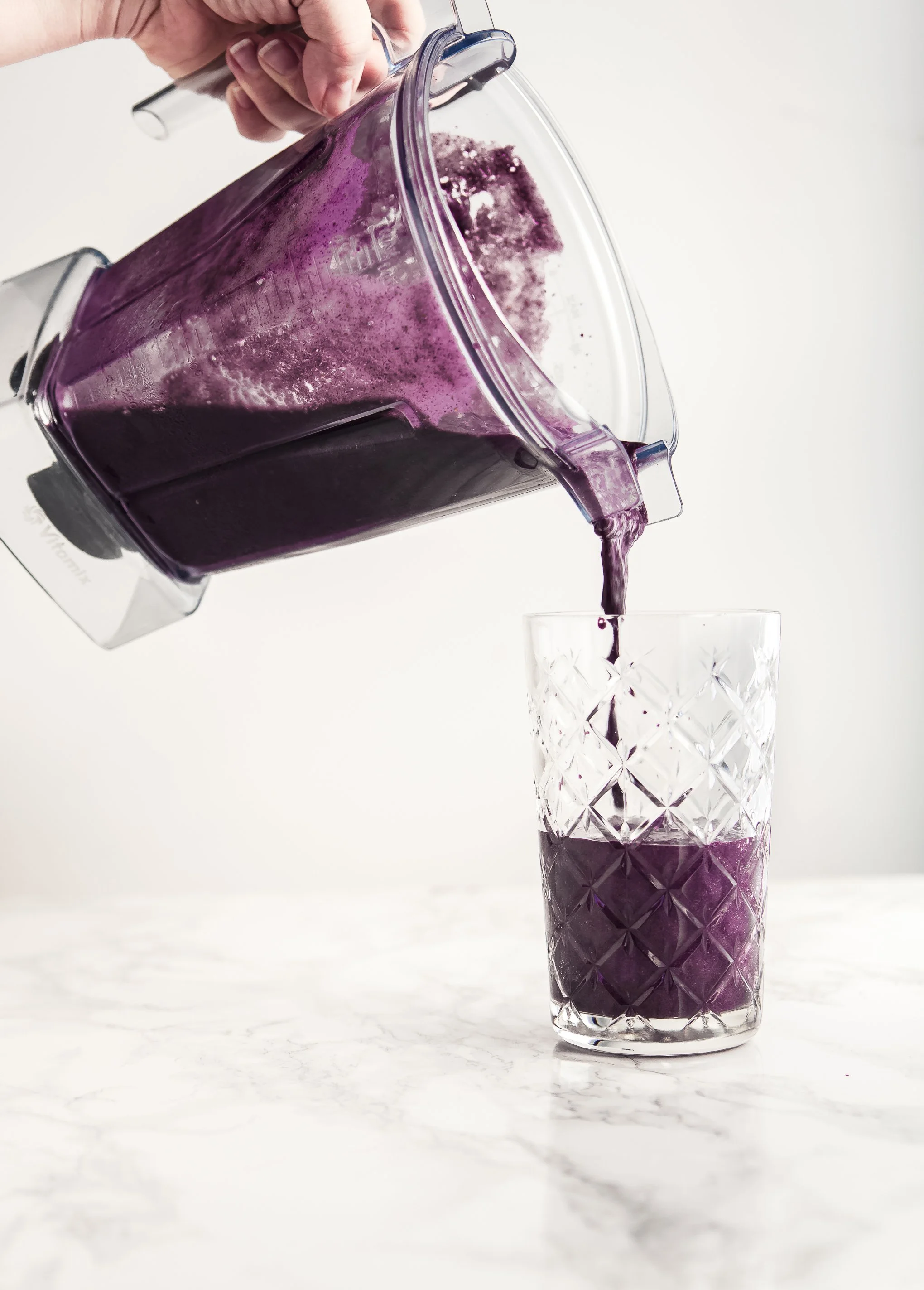 A person pouring purple smoothie from a blender into a glass on a white marble surface.