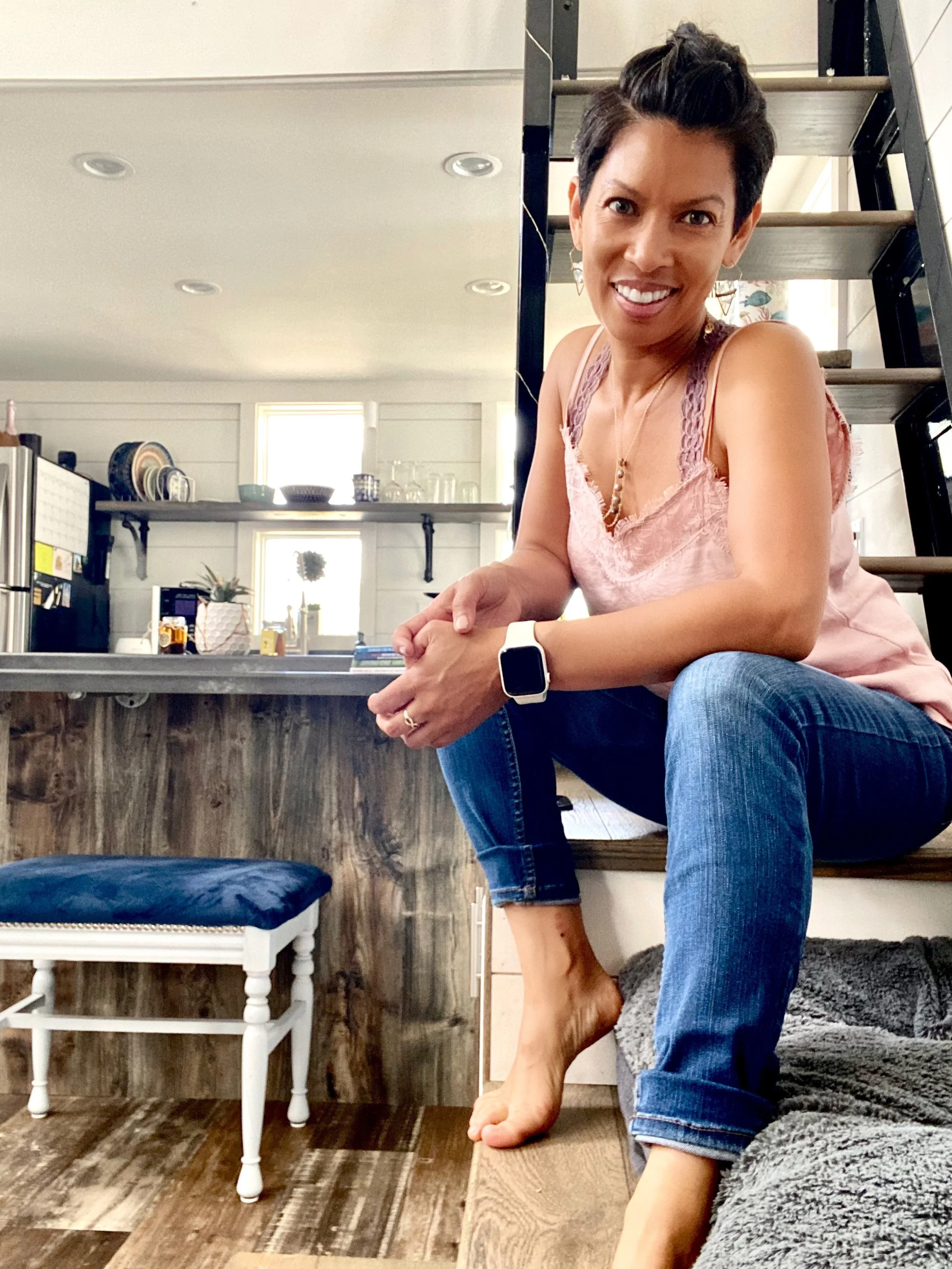 A woman with short dark hair smiling, sitting on a wooden ledge indoors, wearing a pink sleeveless top, blue jeans, and a smartwatch. Behind her is a kitchen with open shelving and a ladder.