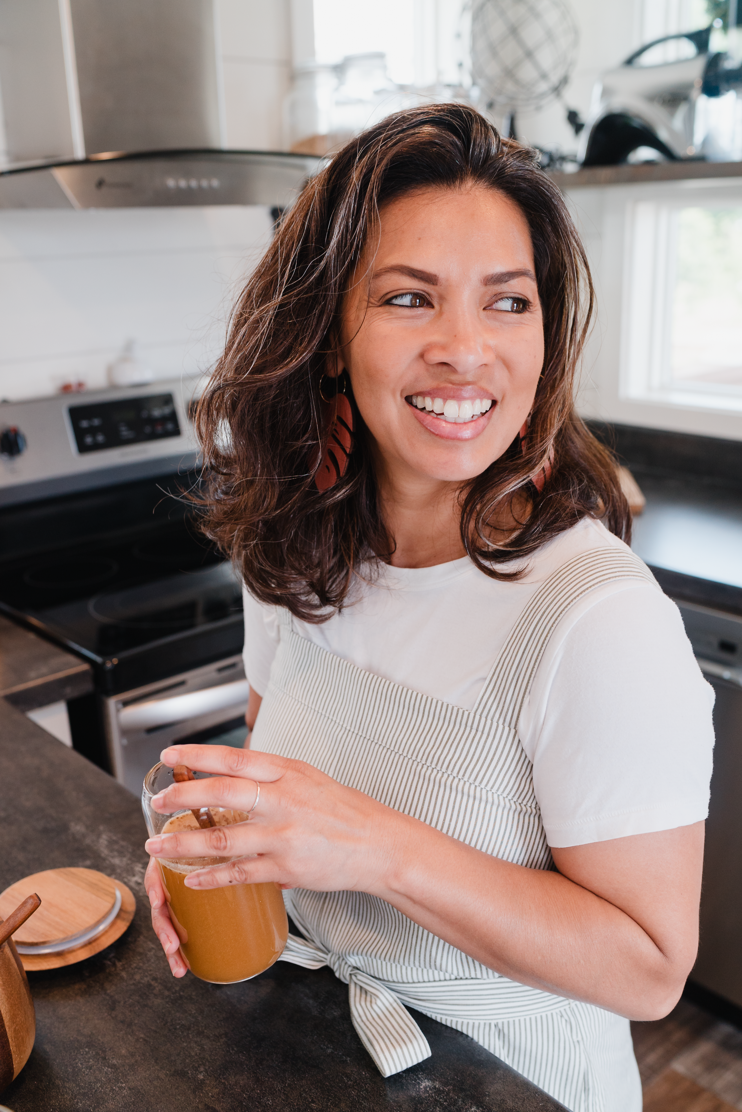 A woman with wavy brown hair and earrings, smiling and looking to her right, holding a glass of iced coffee or latte in a kitchen with a stove, window, and various kitchen items behind her.