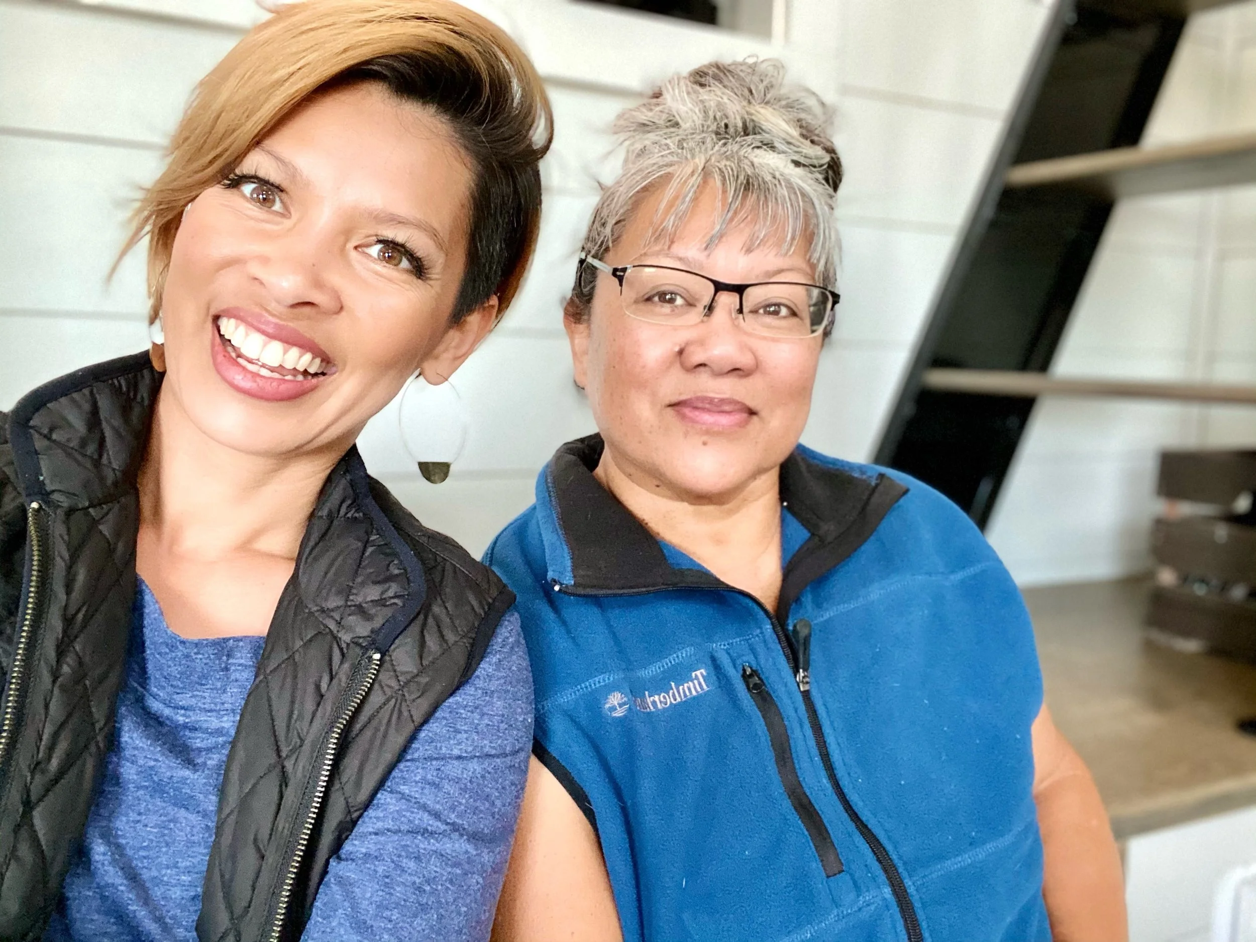 Two women smiling for a selfie indoors. The woman on the left has short hair and is wearing a black vest over a blue shirt. The woman on the right has gray hair, glasses, and is wearing a blue fleece vest. They are sitting near a white wall with shelves behind them.