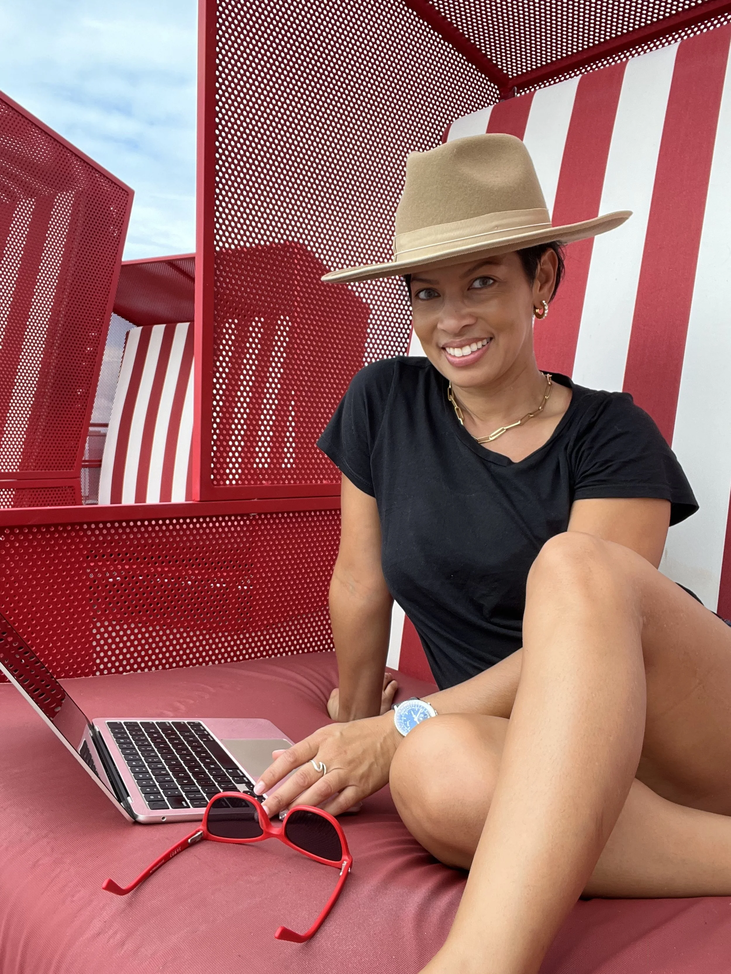 Woman with short hair and gold jewelry, wearing a beige hat and black t-shirt, sitting on a pink cushioned surface with a laptop and red sunglasses, against a red and white striped background.
