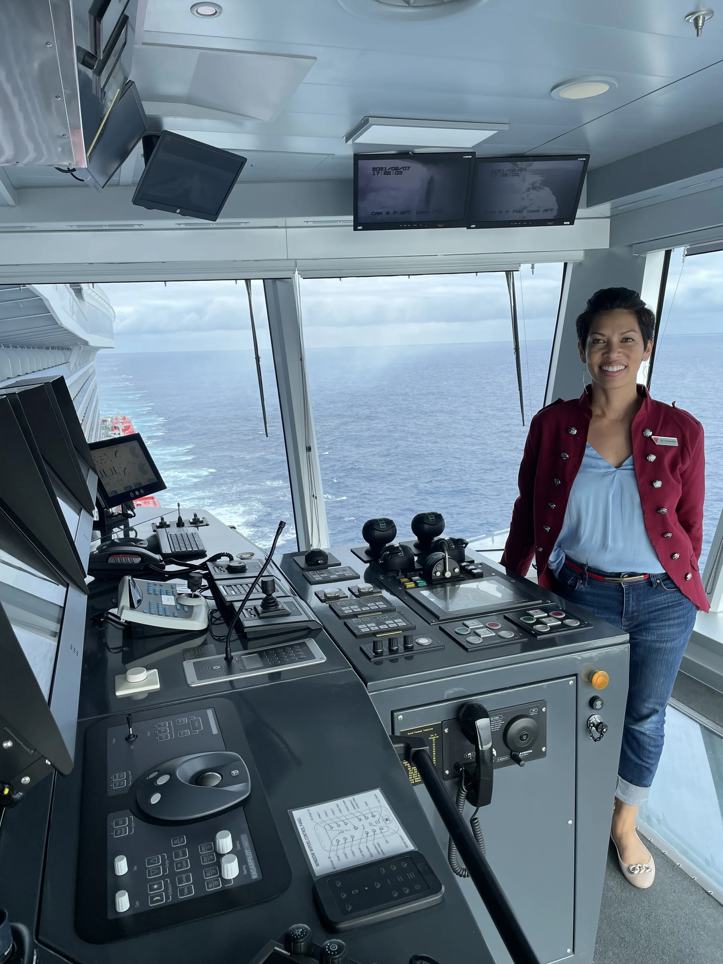 A smiling woman standing in the bridge of a cruise ship, with control panels, monitors, and the ocean visible through large windows behind her.