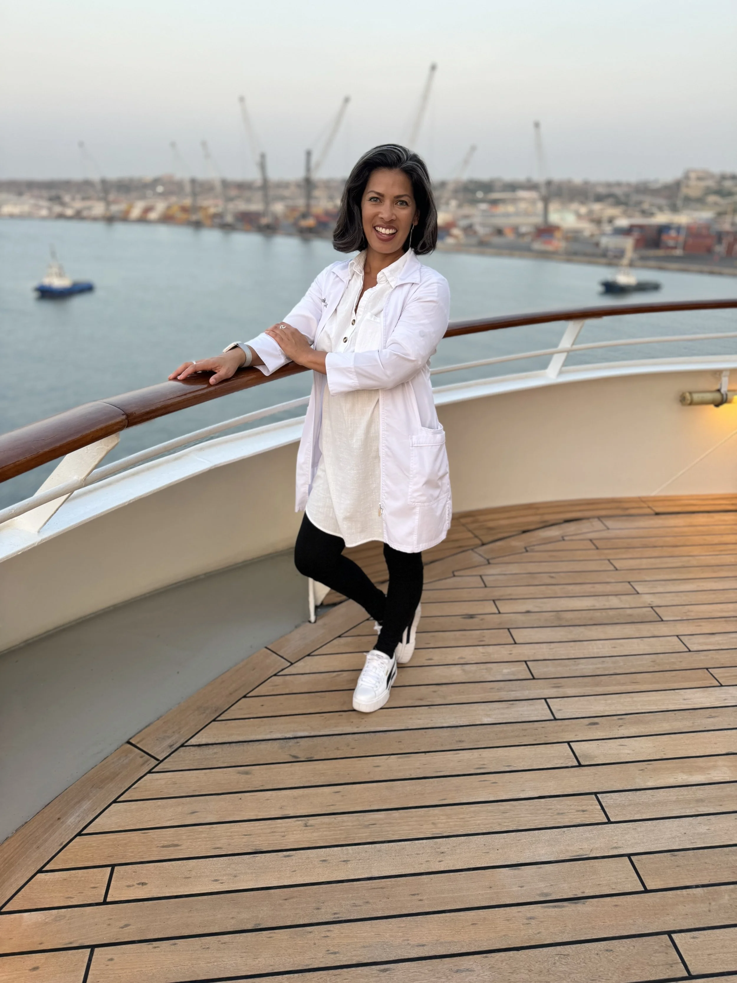A woman standing on the deck of a ship, smiling at the camera with a harbor and cranes in the background.