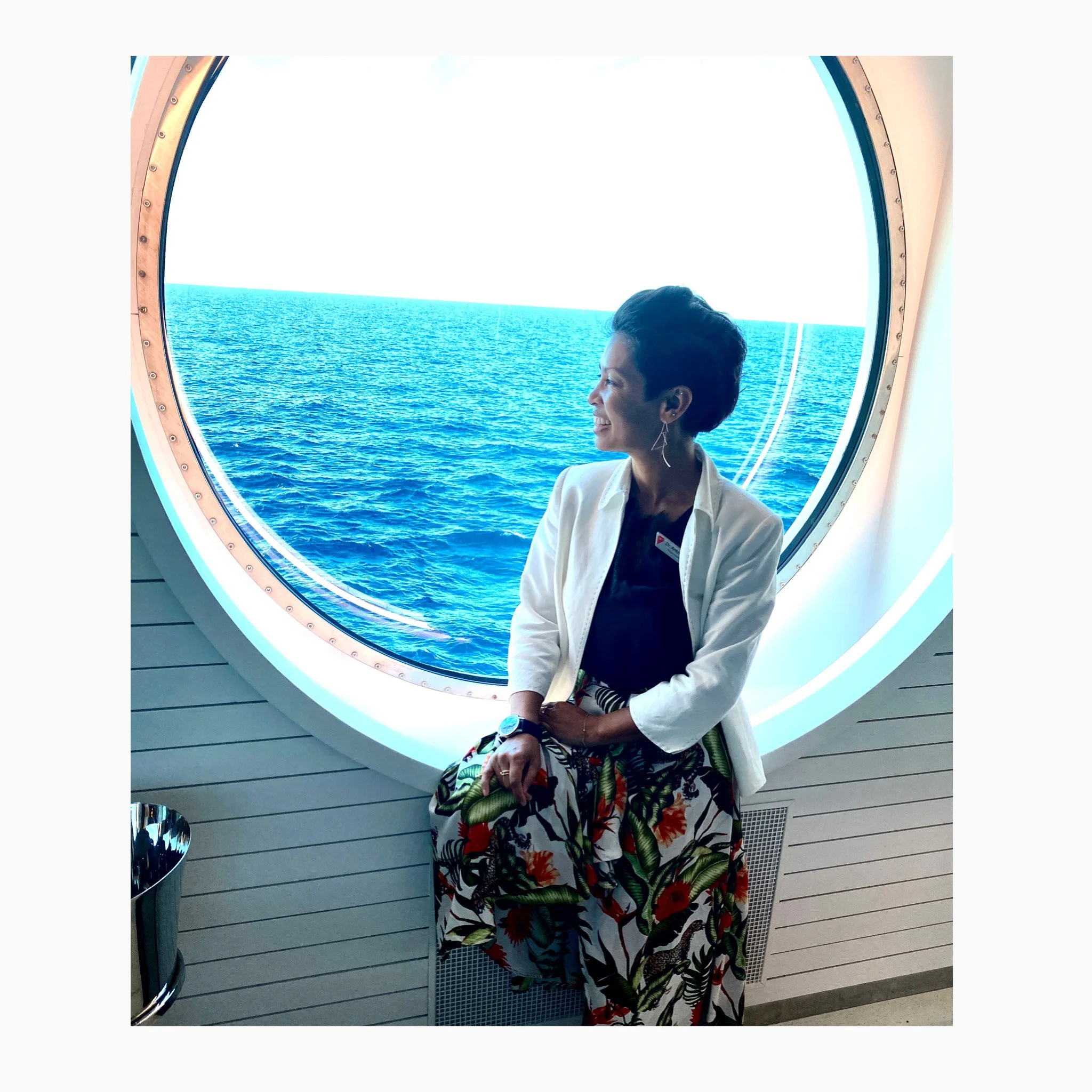 A woman sitting by a large circular window overlooking the ocean, smiling and looking to the side.