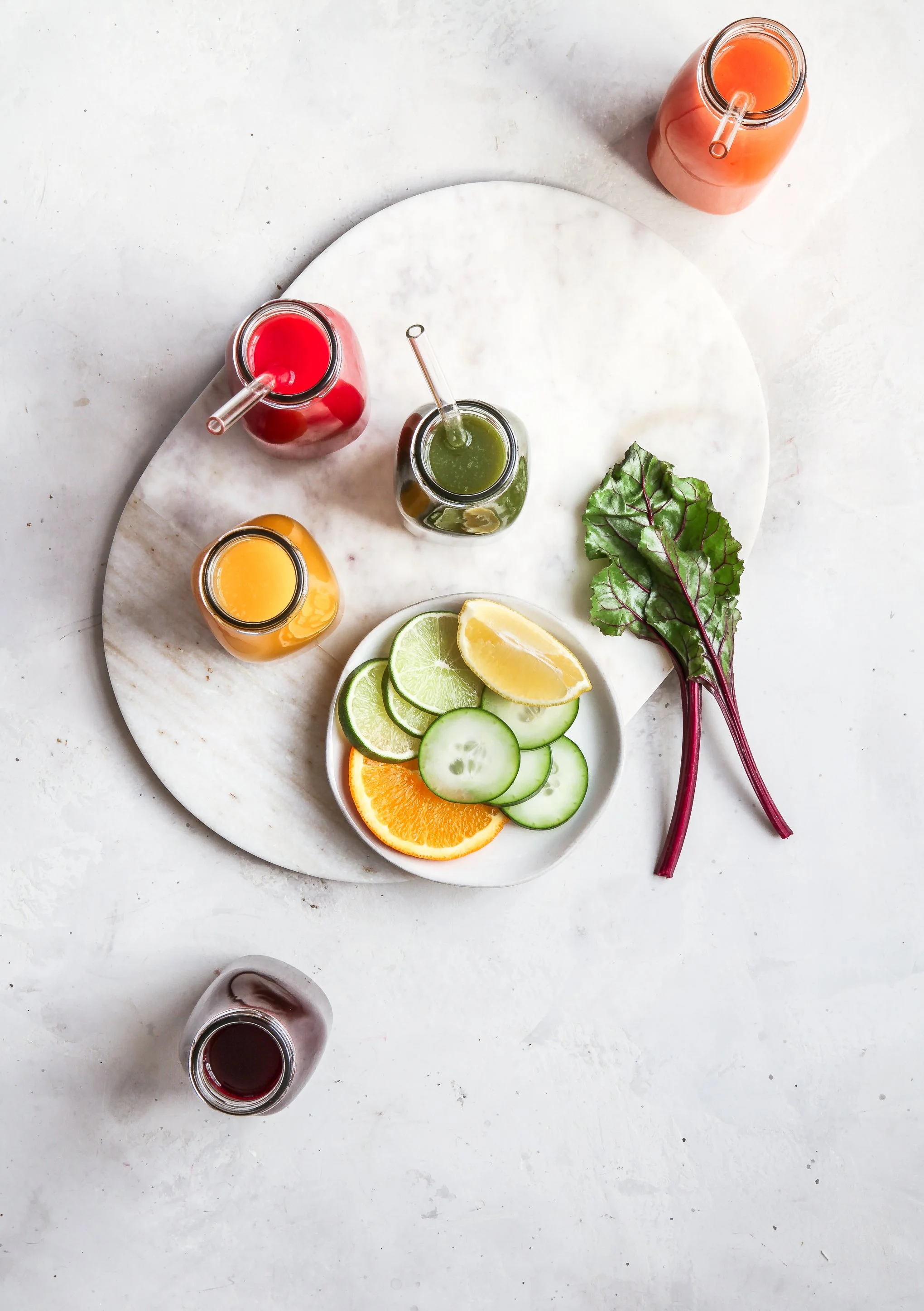 A white marble round board with five small glass bottles containing different colored fruit juices, arranged around a small white plate with citrus slices and cucumber. A leafy green vegetable with purple stems is placed next to the plate. An additional small glass of dark red juice is placed at the bottom of the image on a white surface.