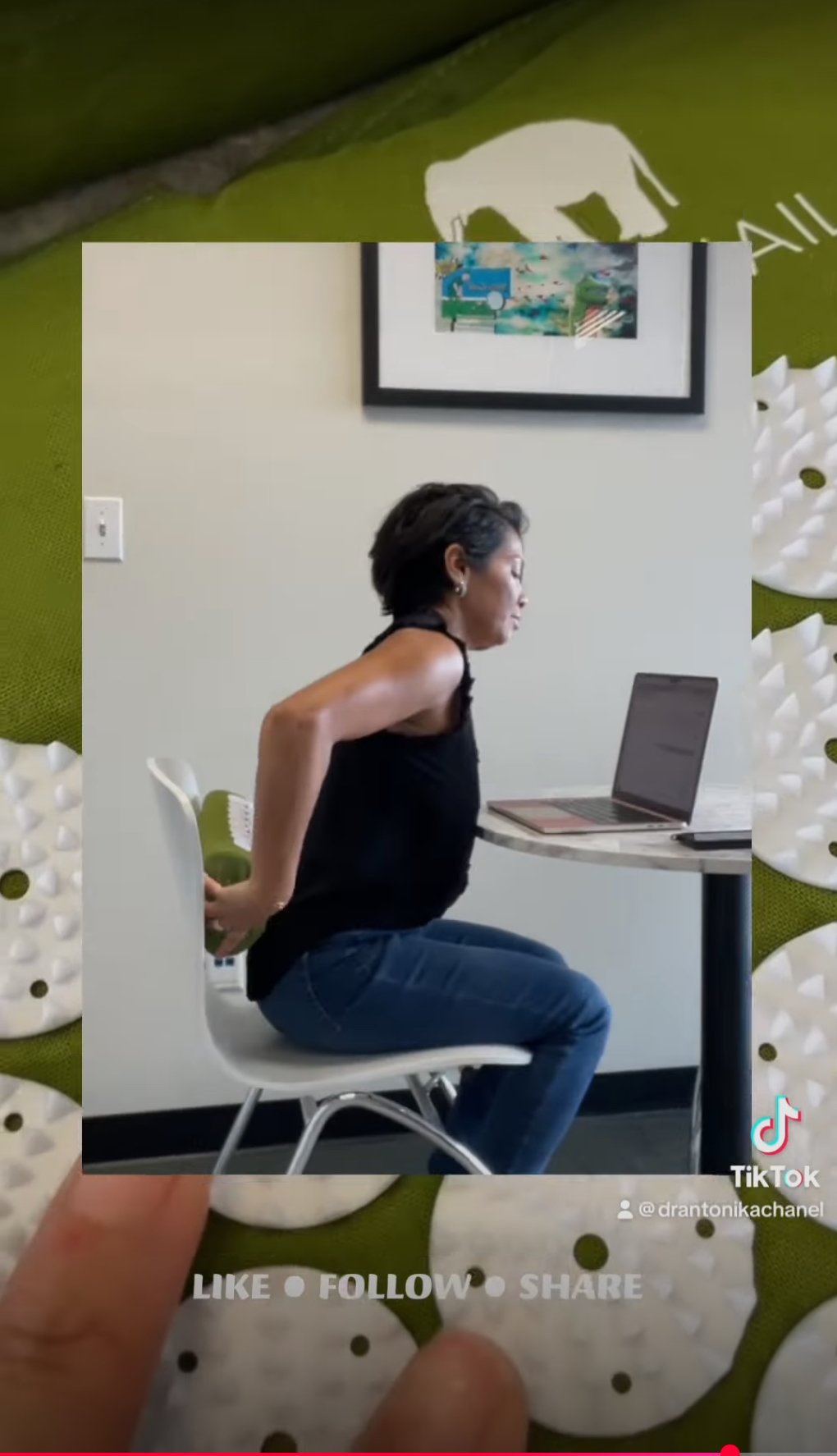 Woman in black tank top and blue jeans sitting on a chair in front of a laptop indoors.