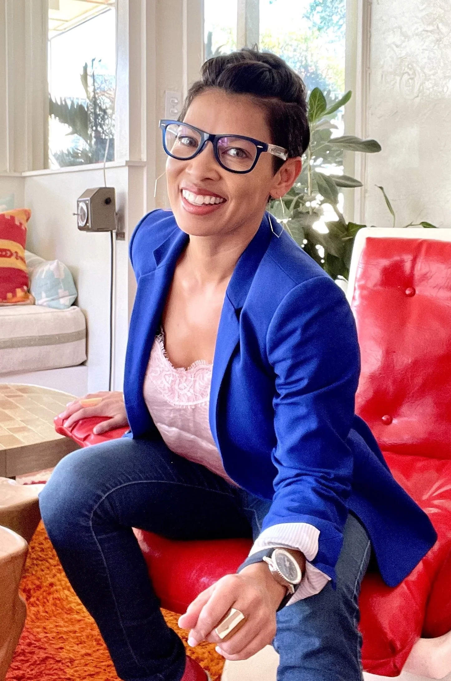 A woman with short dark hair and glasses smiling and sitting on a red chair in a sunlit room with plants and a window.
