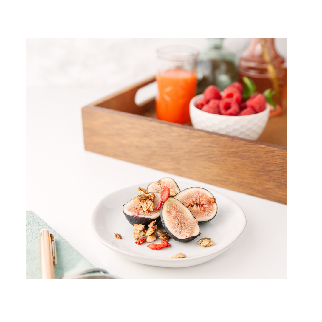 Halved figs topped with granola and goji berries on a white plate in front of a wooden tray with a bowl of raspberries and a glass of orange juice.