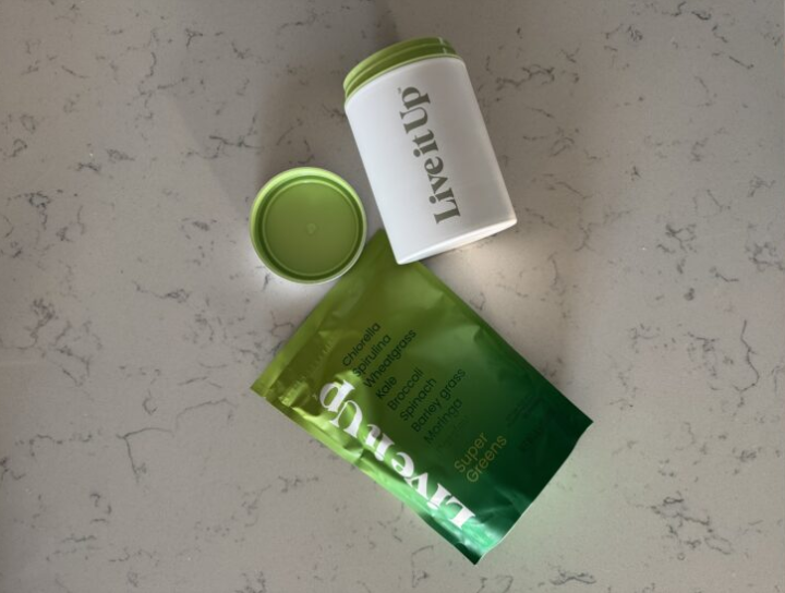 A white and green dietary supplement container with lid, a green bag of LiveUp Super Greens powder, and a green lid on a speckled countertop.