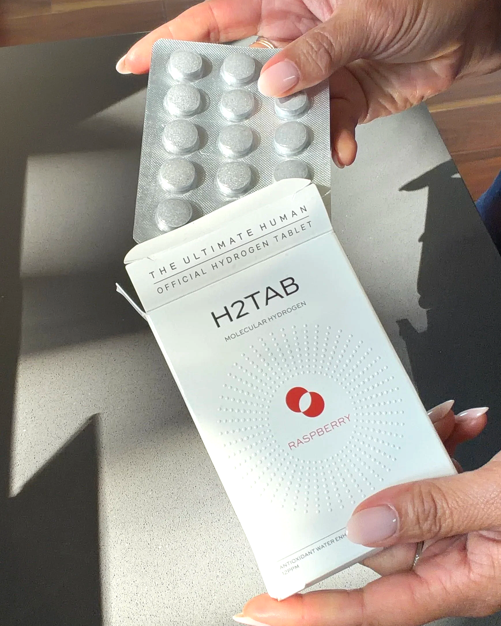 Hand holding a blister pack of hydrogen tablets with a partially visible box labeled 'The Ultimate Human Official Hydrogen Tablet' and 'H2Tab' with raspberry flavor