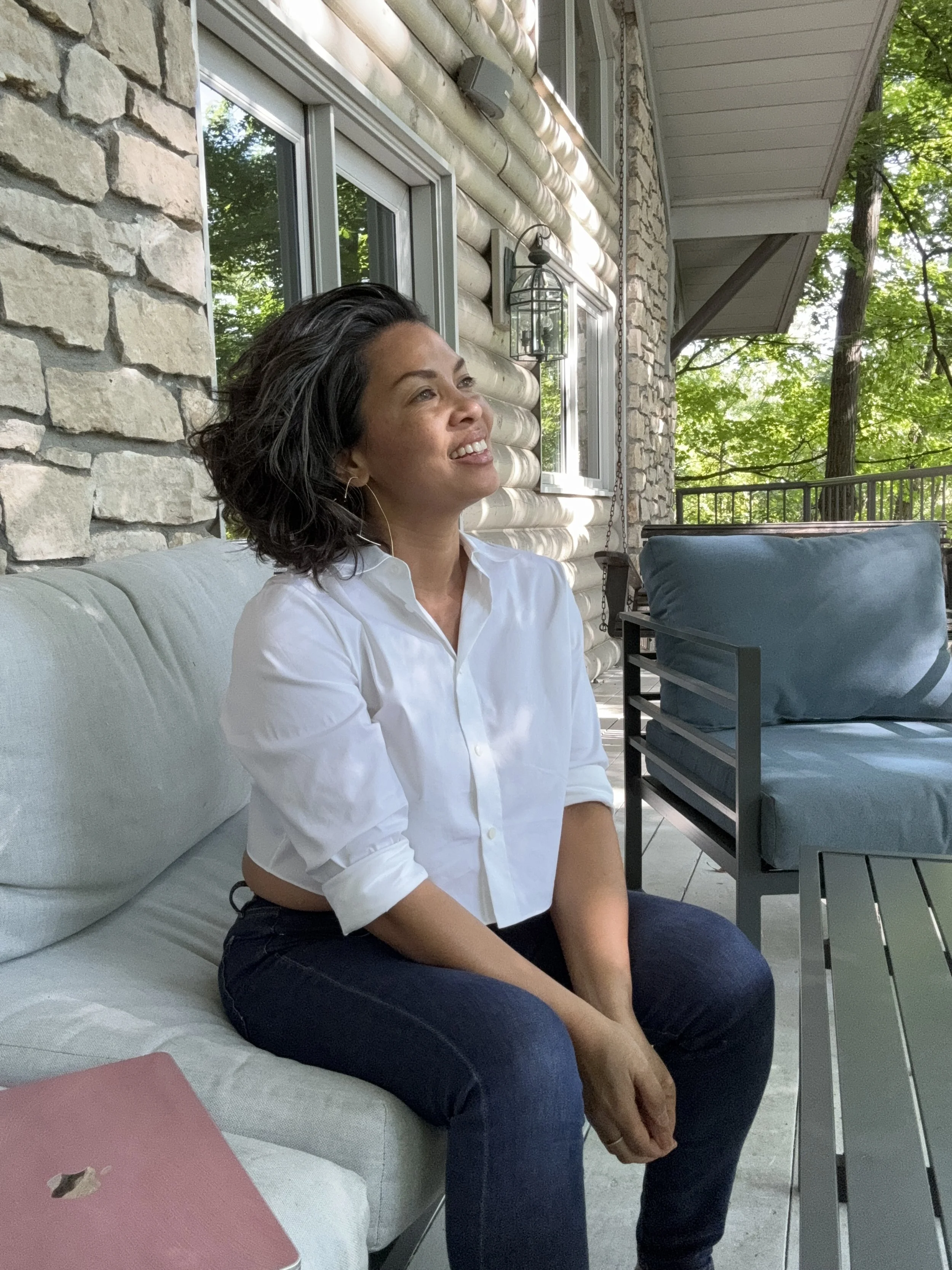 Woman sitting on outdoor patio sofa, smiling and looking to the side, with trees and a house exterior in the background.