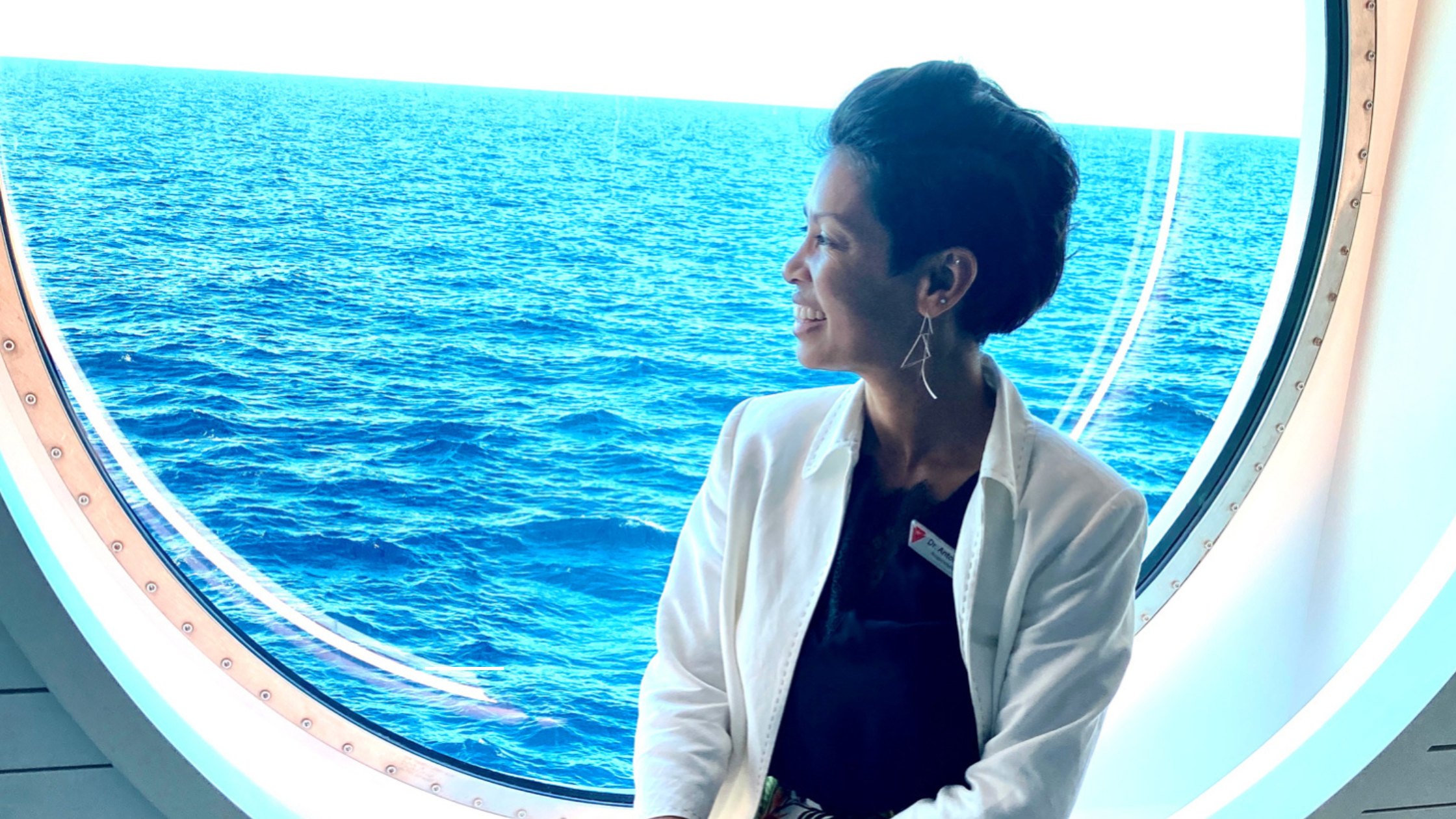 Woman with short dark hair smiling and looking outside through a large, round window at the blue ocean.