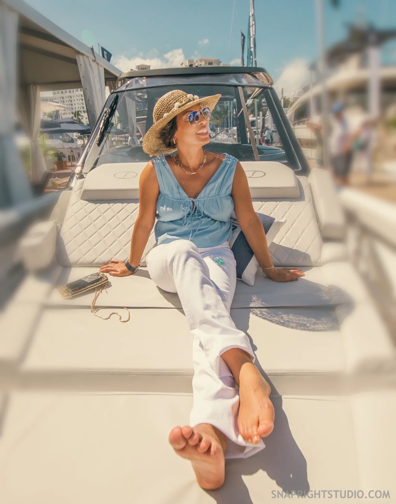 Woman sitting on the deck of a luxury yacht, wearing a straw hat, sunglasses, a light blue top, and white pants, with marina and other boats in the background.