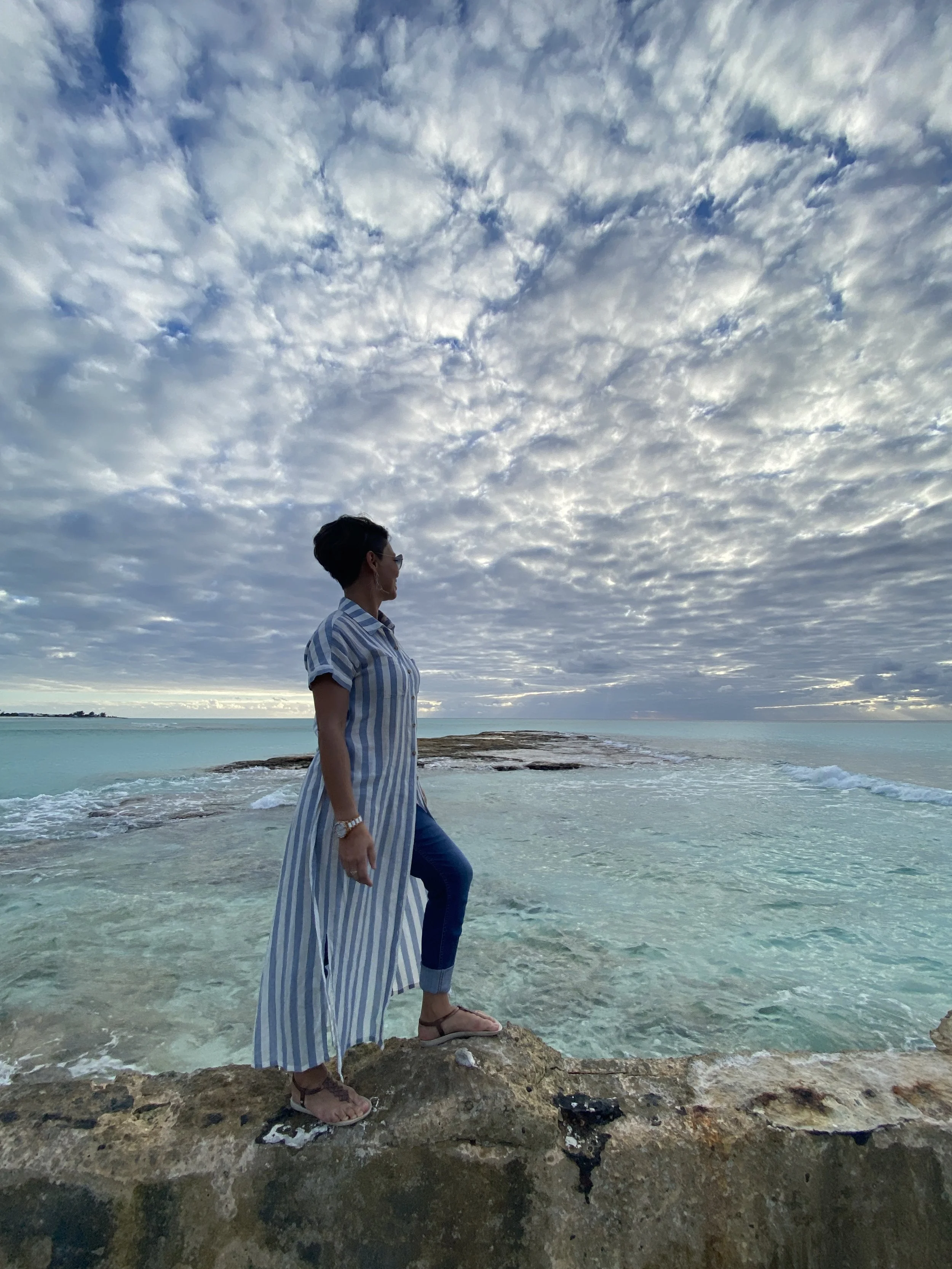 Woman standing on rocks by the ocean, wearing a striped long shirt and jeans, looking at the sea with cloudy sky above.