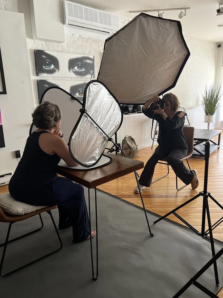 A woman in black clothing takes a portrait of another woman sitting at a table with a white reflector in front of her, while a photographer uses a camera with a large umbrella-style softbox attached. The setting appears to be a photography studio or a well-lit room with white brick walls, artwork, and a potted plant in the background.