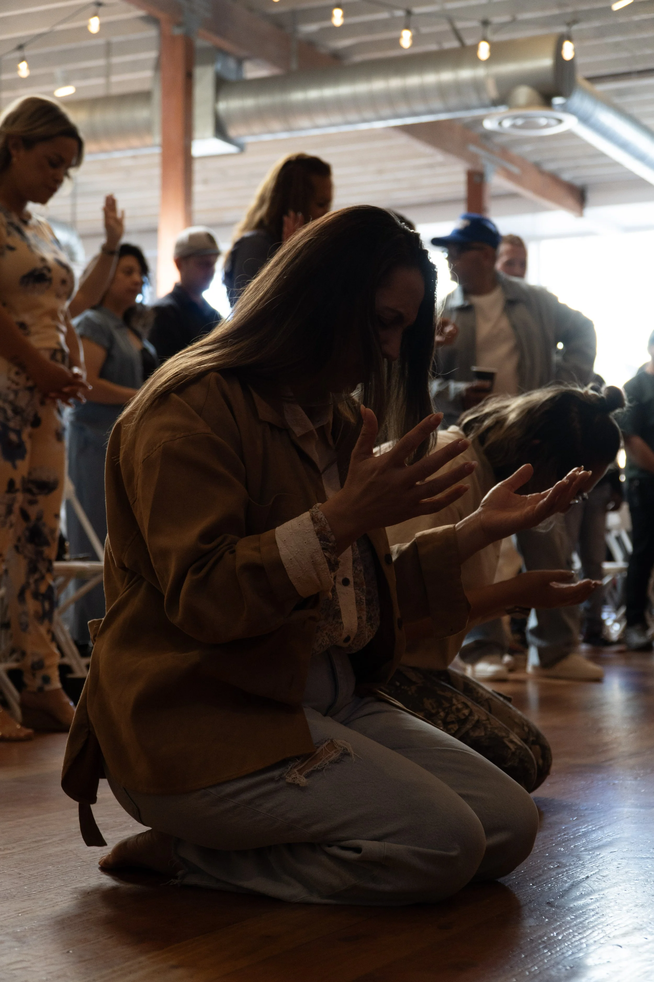 A woman kneeling on the floor with her eyes closed and hands raised, participating in a prayer or meditation, surrounded by other people in the background in a dimly lit room with wooden floors and exposed ceiling beams.