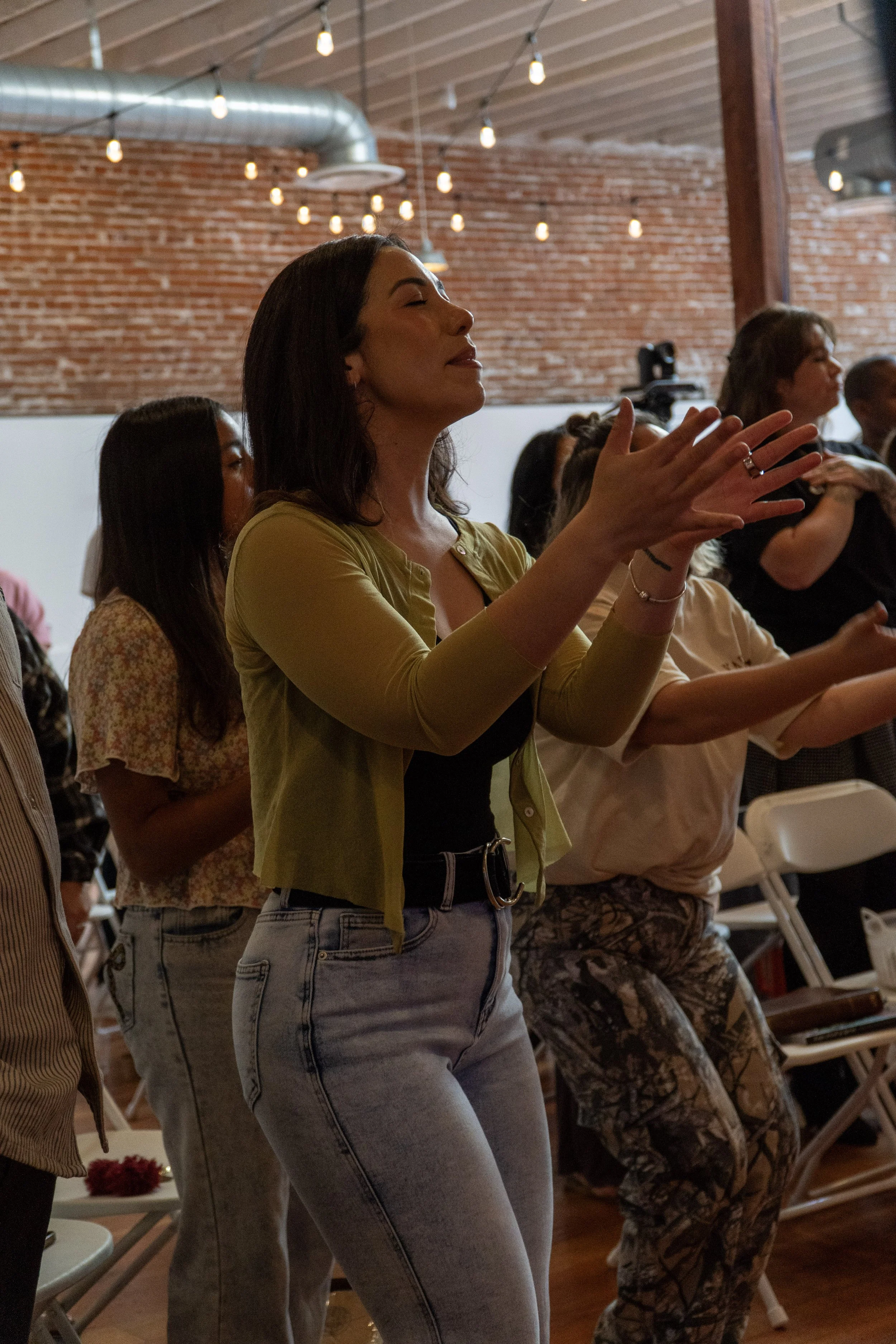 A woman with dark hair and light skin, wearing a yellow jacket and high-waisted jeans, is standing with her eyes closed and hands raised and clasped, participating in a group activity or prayer in a rustic indoor setting with brick walls and string l